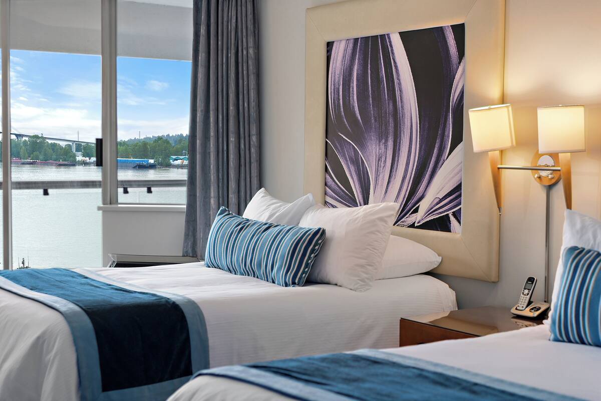 A hotel room with a large window, a bed with blue and white bedding, a wall-mounted lamp, and a painting of leaves.