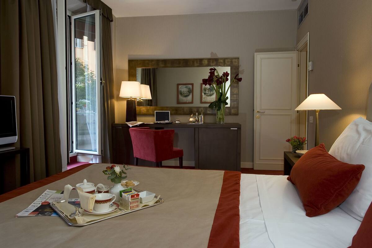 A hotel room with a bed, a desk, a chair, a television, and a dining table with breakfast items.