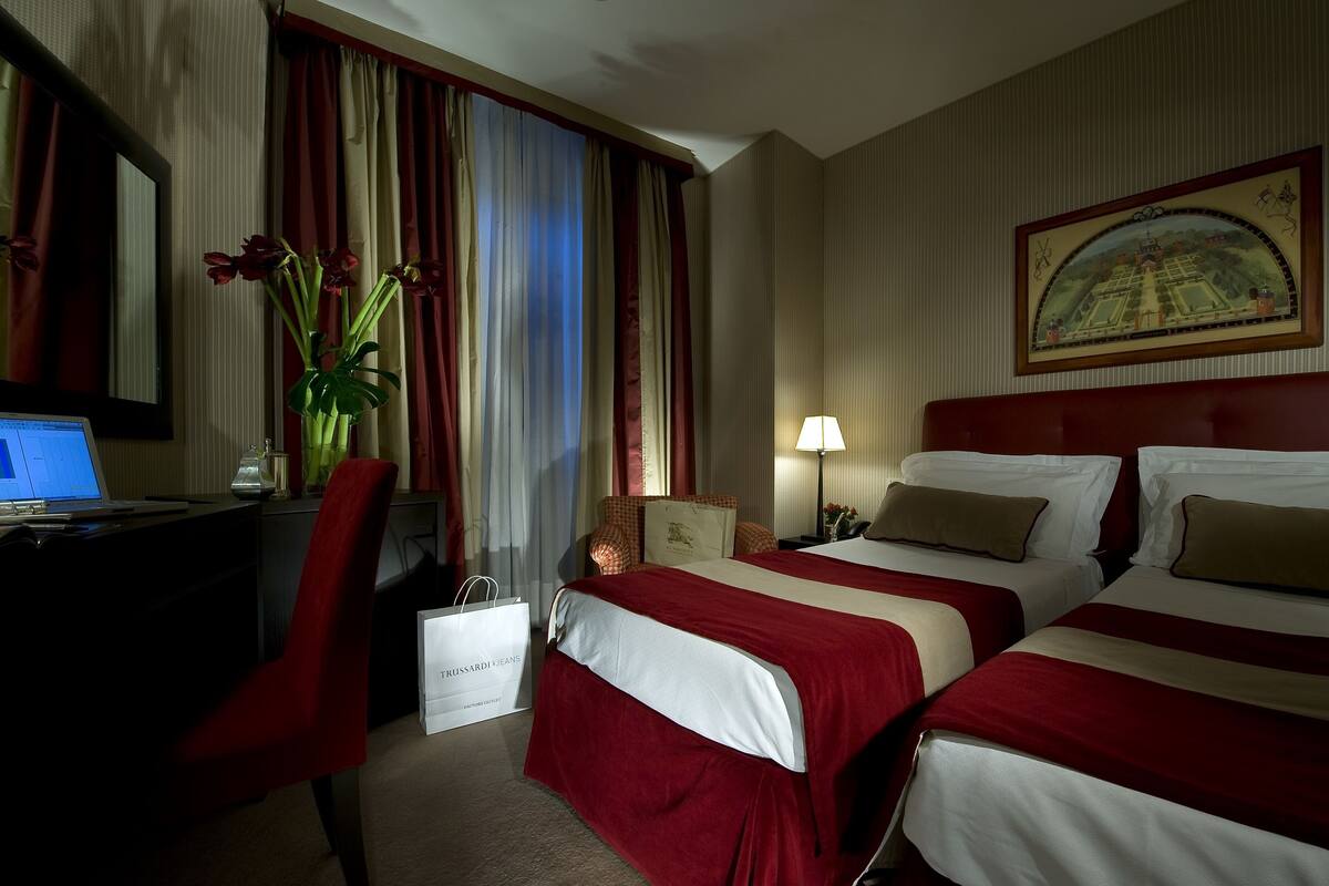 A hotel room with two beds, a desk with a laptop, a vase with flowers, and a painting on the wall.