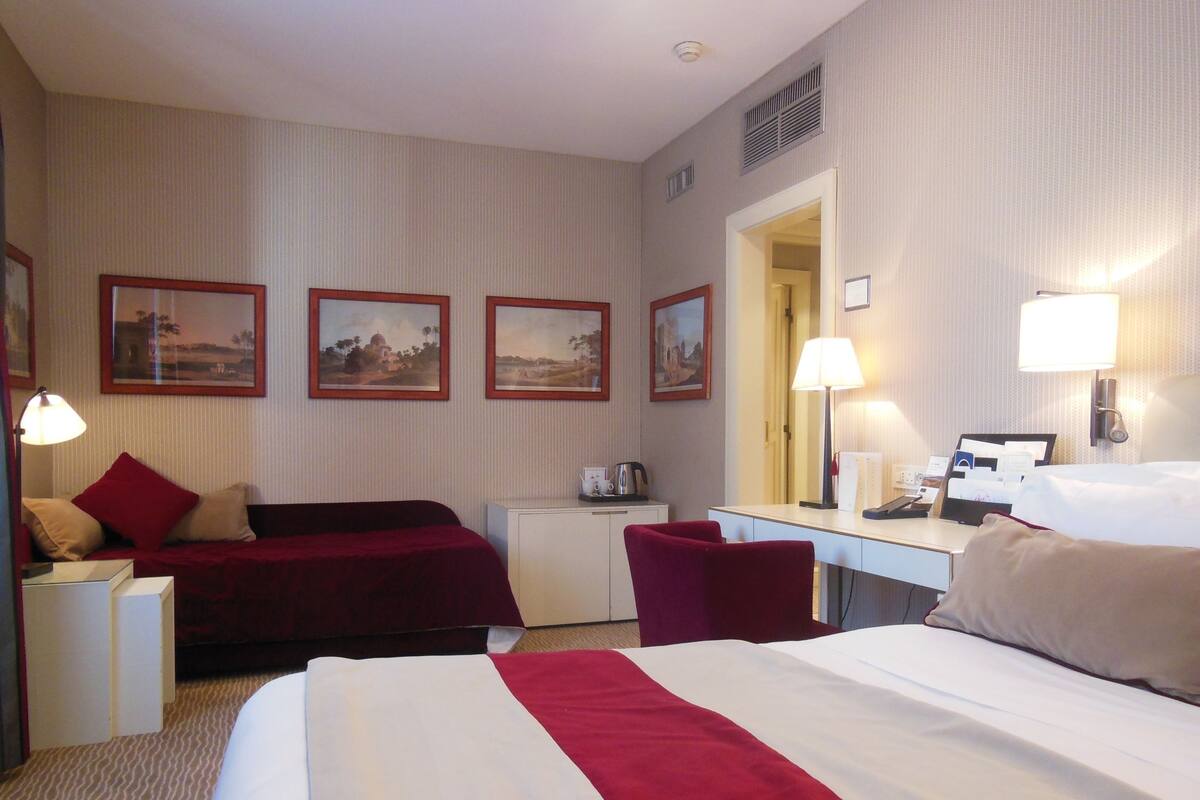 A hotel room with a bed, a desk, a chair, a small table, and three framed pictures on the wall.