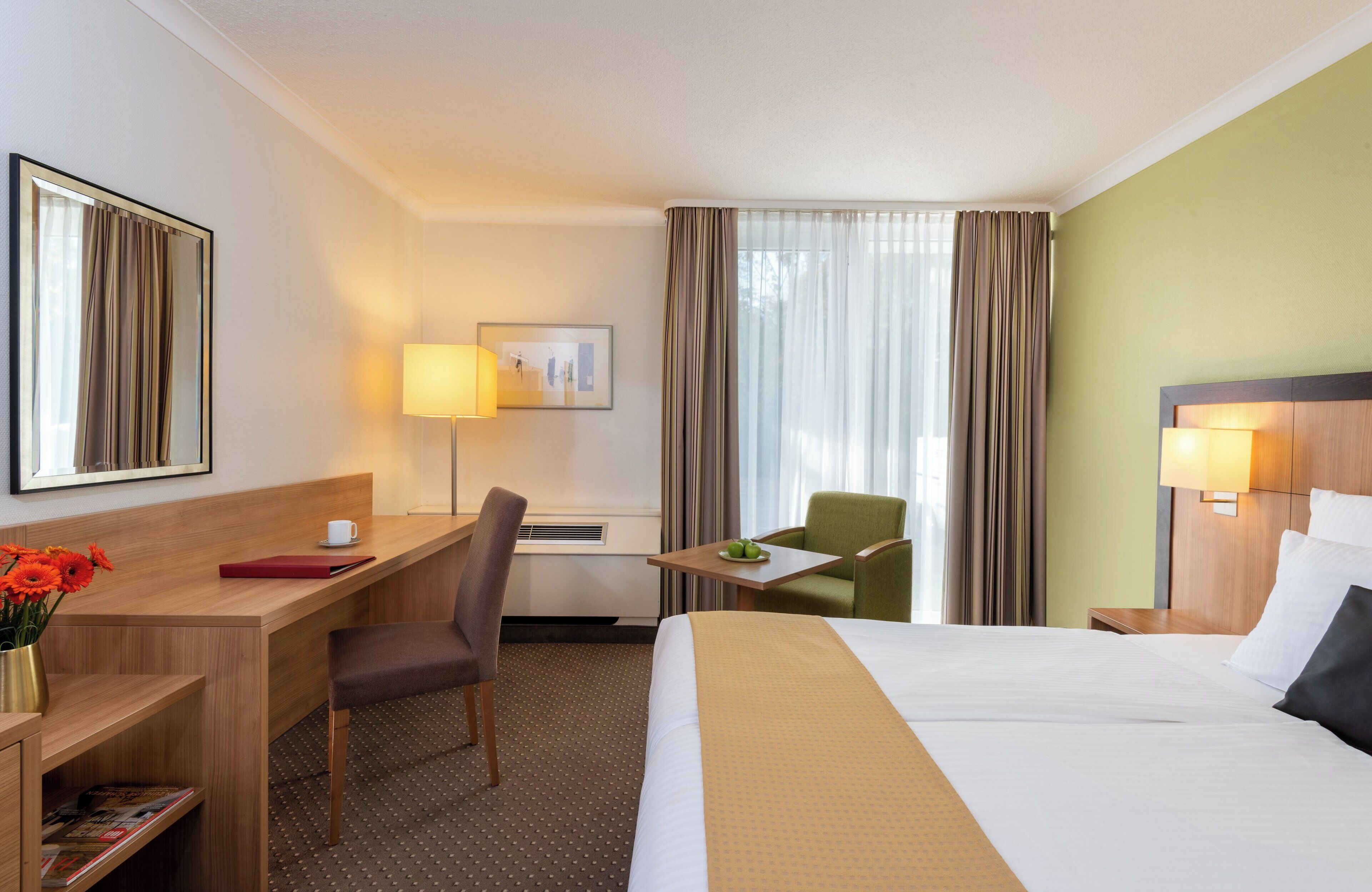 A hotel room with a bed, desk, chair, and a small table with a lamp.