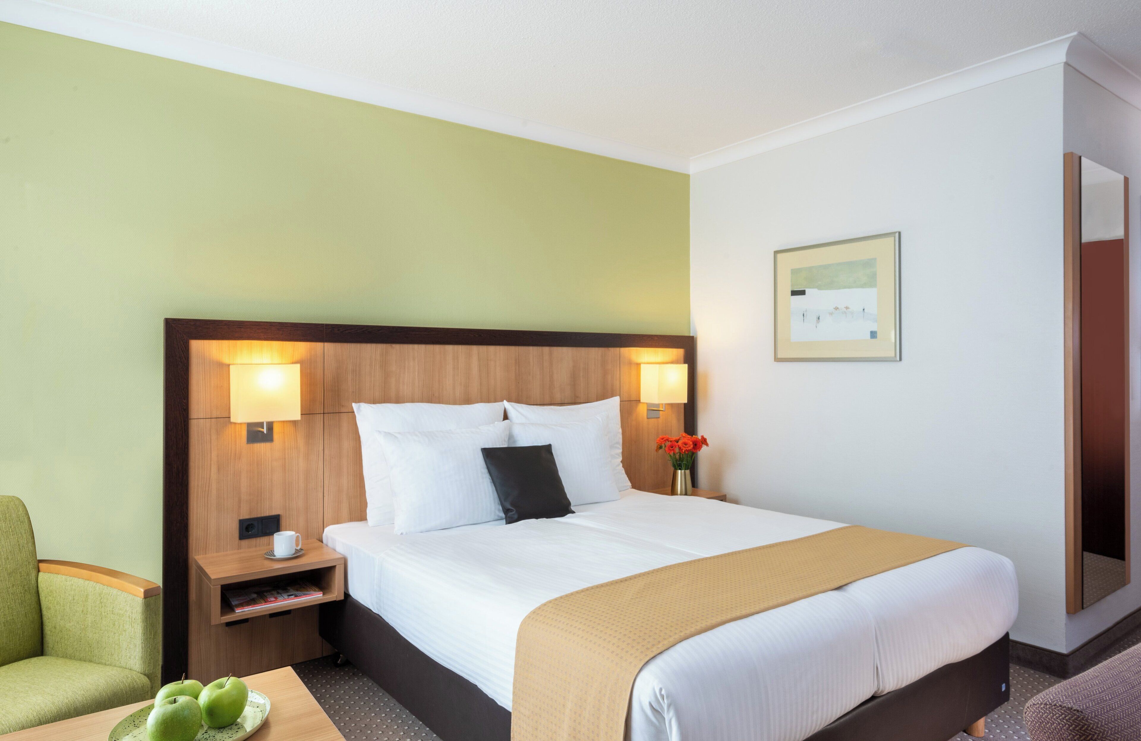 Una habitación de hotel con una cama grande, dos lámparas de noche, una mesita de noche con una taza y una revista, una silla verde y una pequeña mesa con manzanas verdes.