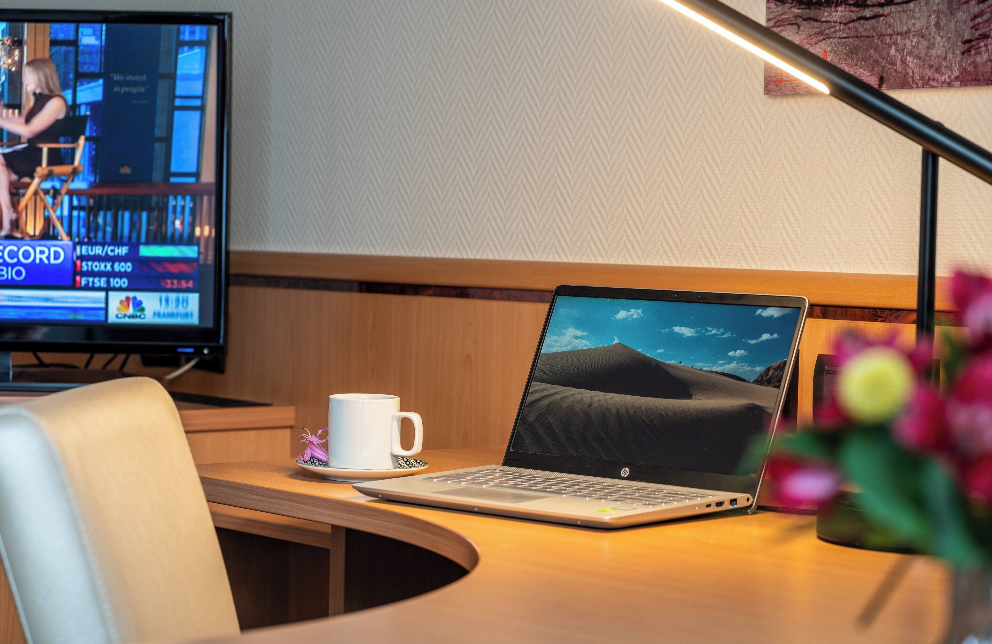 A laptop on a desk with a desert landscape on the screen, a white mug, and a television displaying financial news in the background.