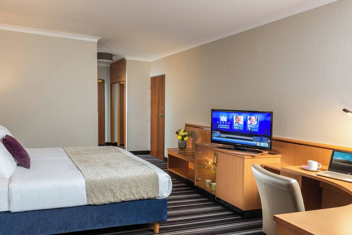 Una habitación de hotel con cama, un escritorio con una computadora portátil, televisión y una mesita con una taza.