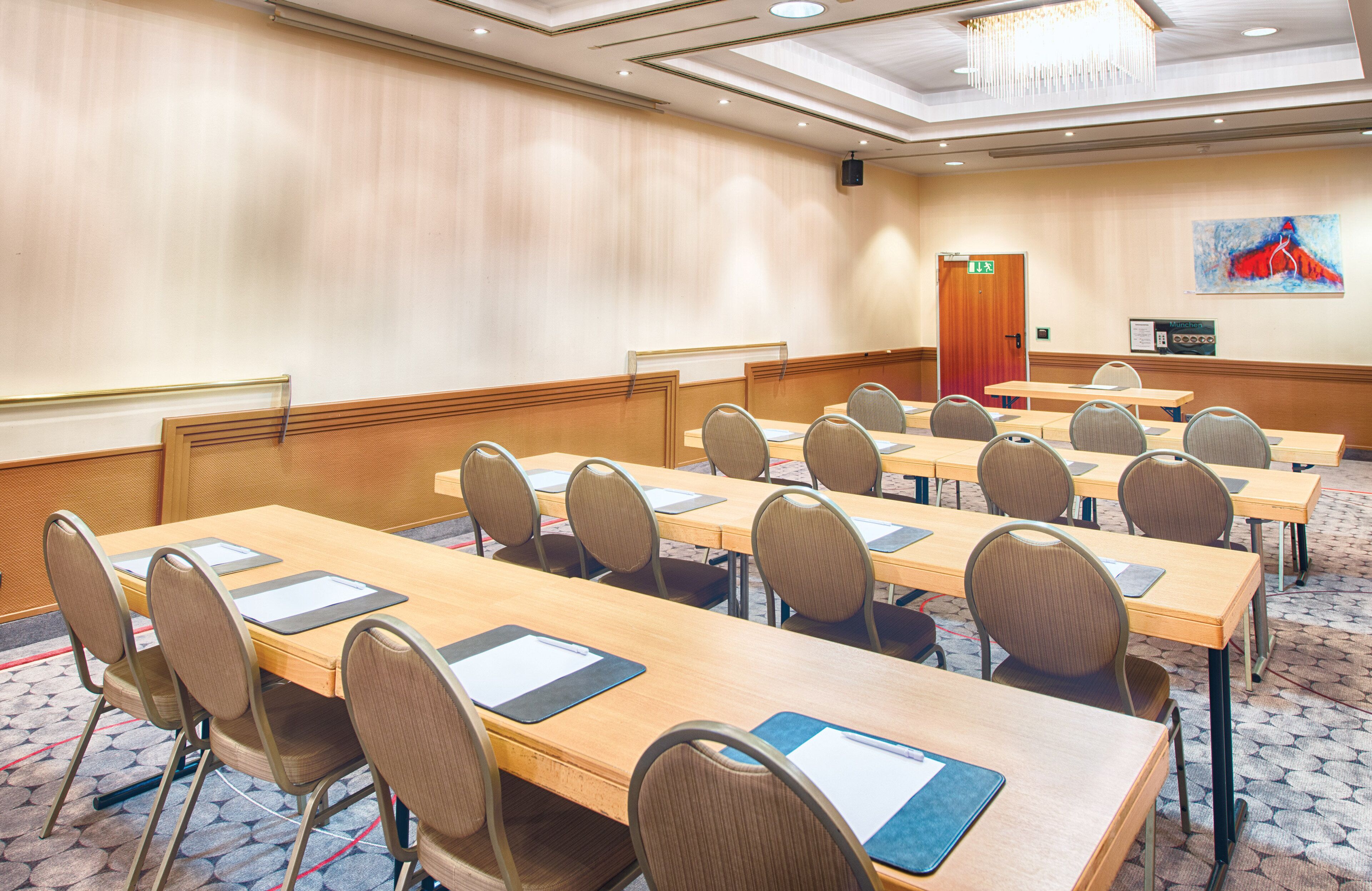 A conference room with multiple tables and chairs, a wooden panel wall, a chandelier, and a door with an exit sign.