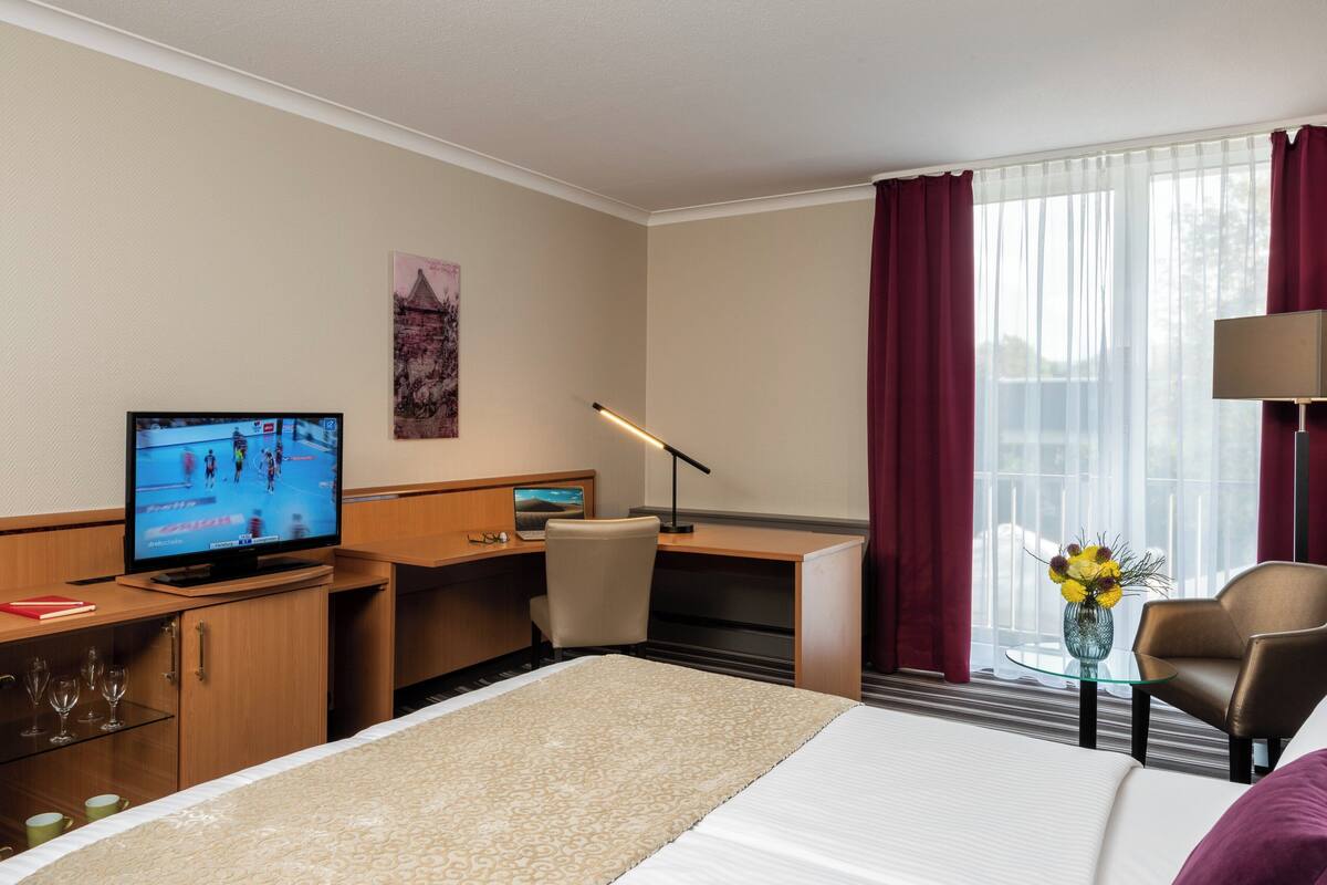 Habitación de hotel con cama, escritorio con computadora, televisor, silla, lámpara, jarrón con flores y ventana con cortinas.