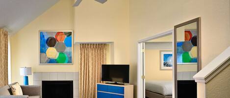 Habitación de hotel con sofá, mesa de centro, televisor, chimenea, ventilador de techo y obras de arte en las paredes.