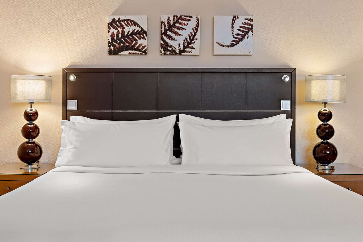 Una cama bien tendida con sábanas blancas, flanqueada por dos mesitas de noche con lámparas, y un cabecero con diseño de cuadrícula.