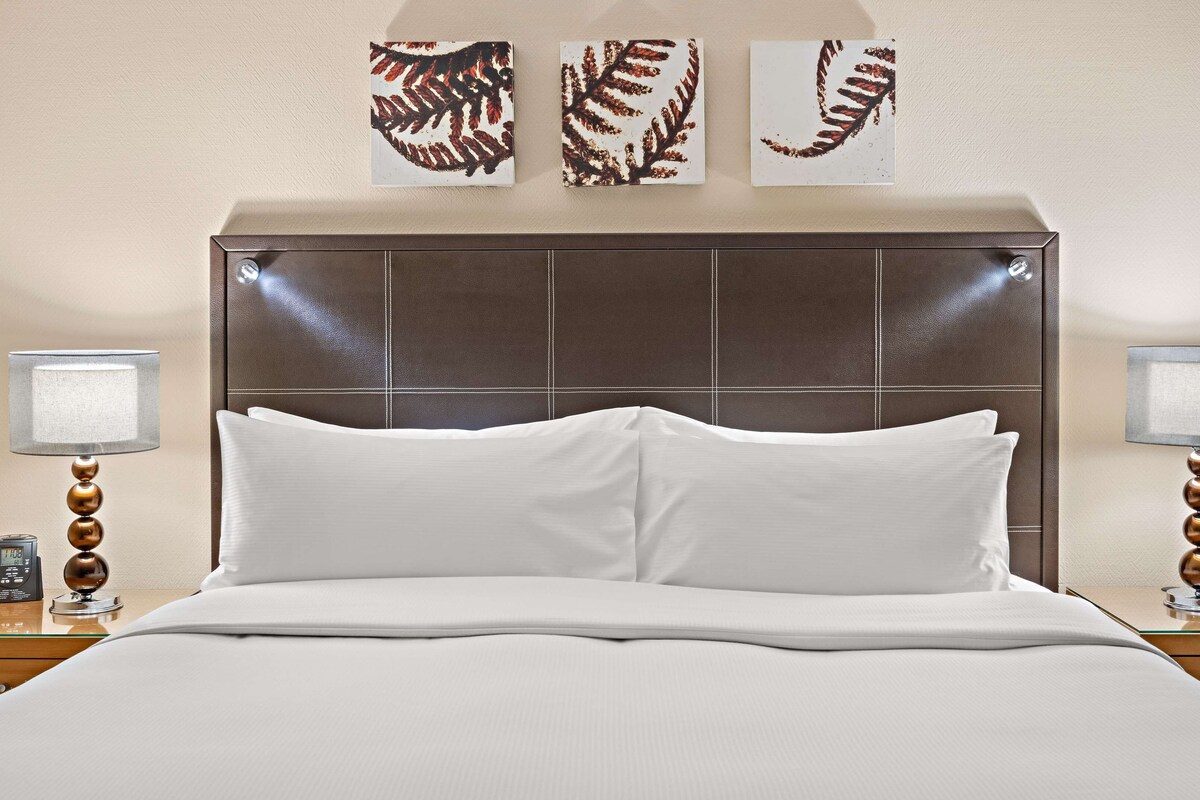 Una cama bien hecha con sábanas blancas, cabecera acolchada y dos lámparas de noche con base de madera.