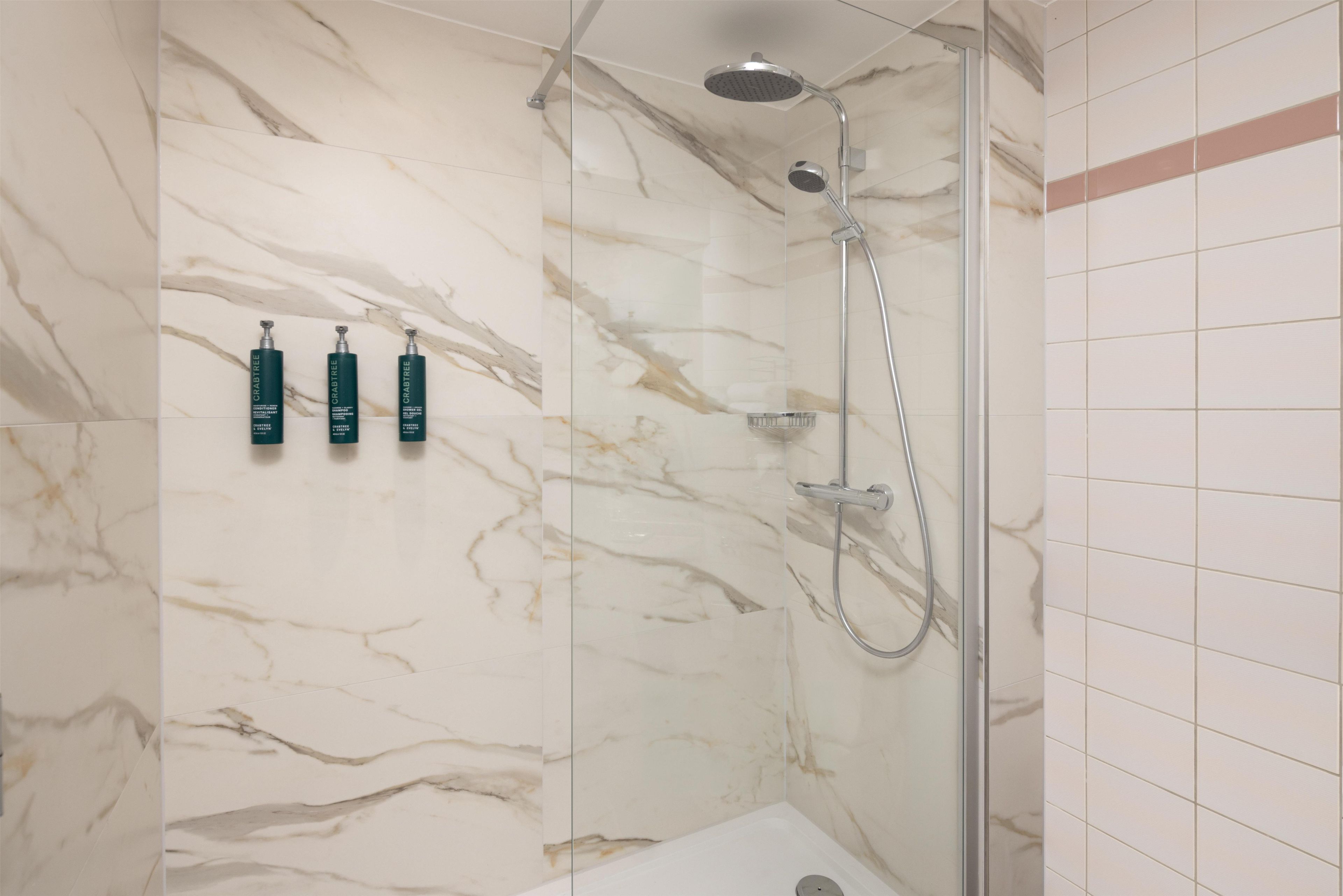 A shower with a handheld showerhead and a wall-mounted faucet, a glass-enclosed shower stall, and three bottles of Crabtree shampoo on the wall.