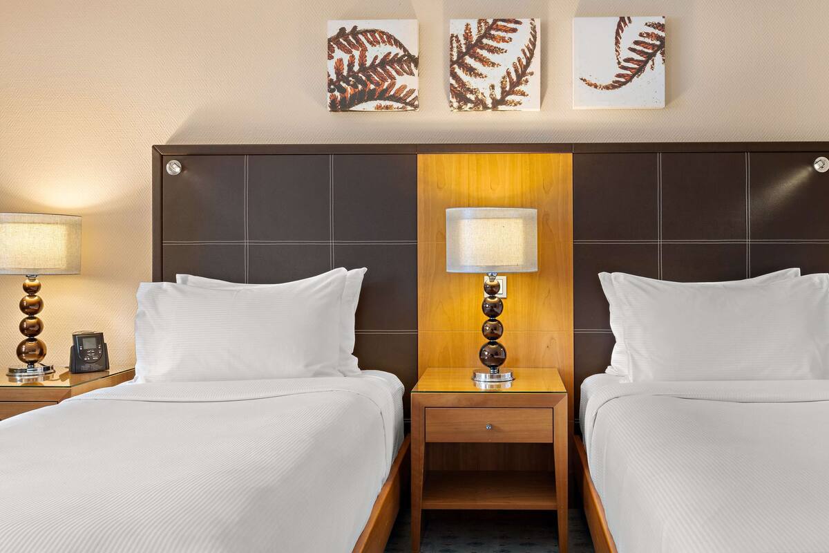 A hotel room with two beds, a nightstand with a lamp, a phone, and artwork on the wall.