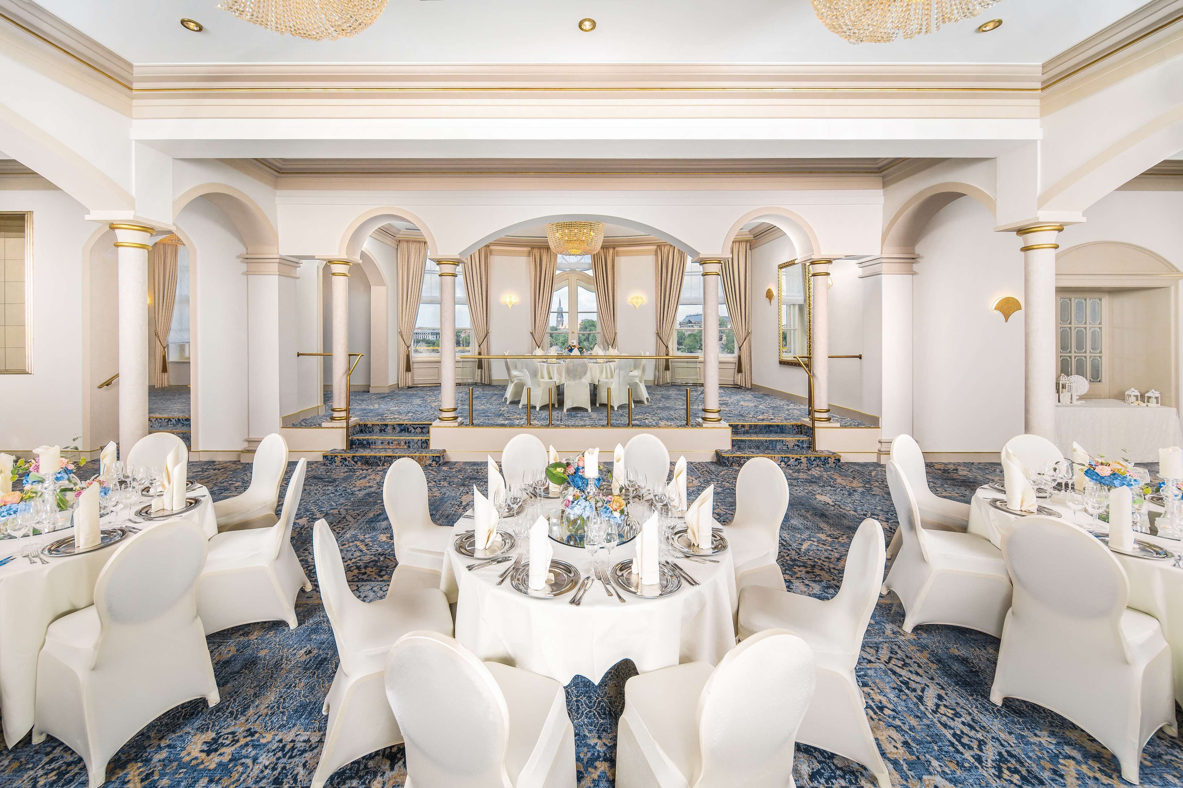 A luxurious banquet hall with white chairs and tables, a blue carpet, and a chandelier.