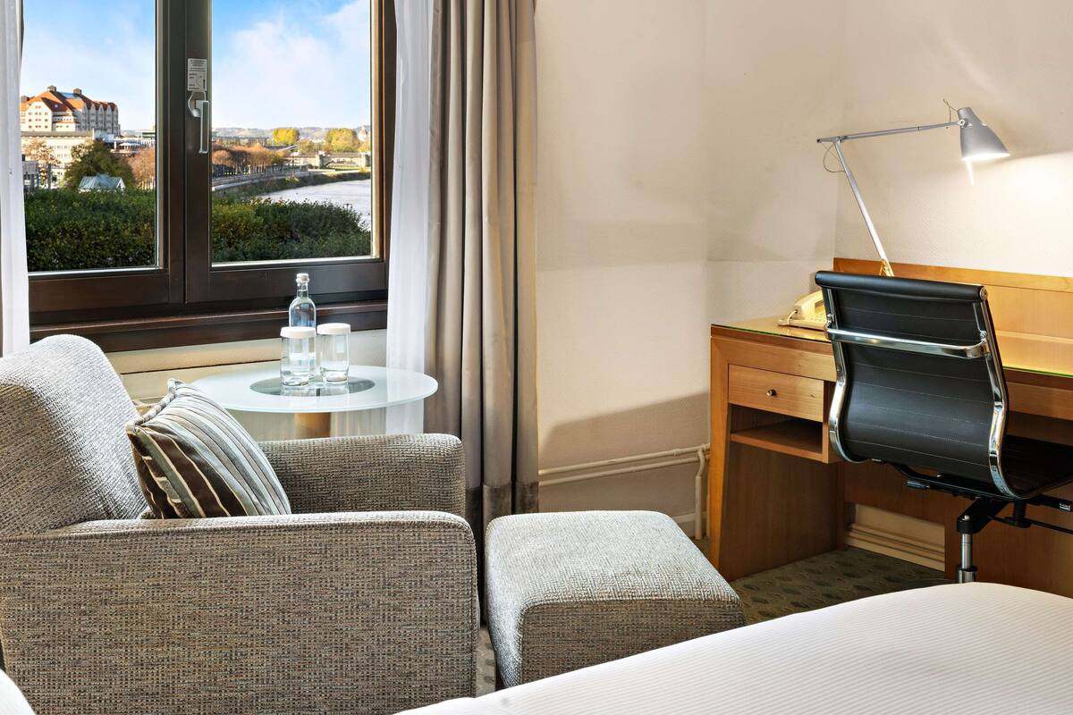 A hotel room with a bed, a desk with a chair, a small round table with a bottle and glass, and a window with a view of buildings and greenery.