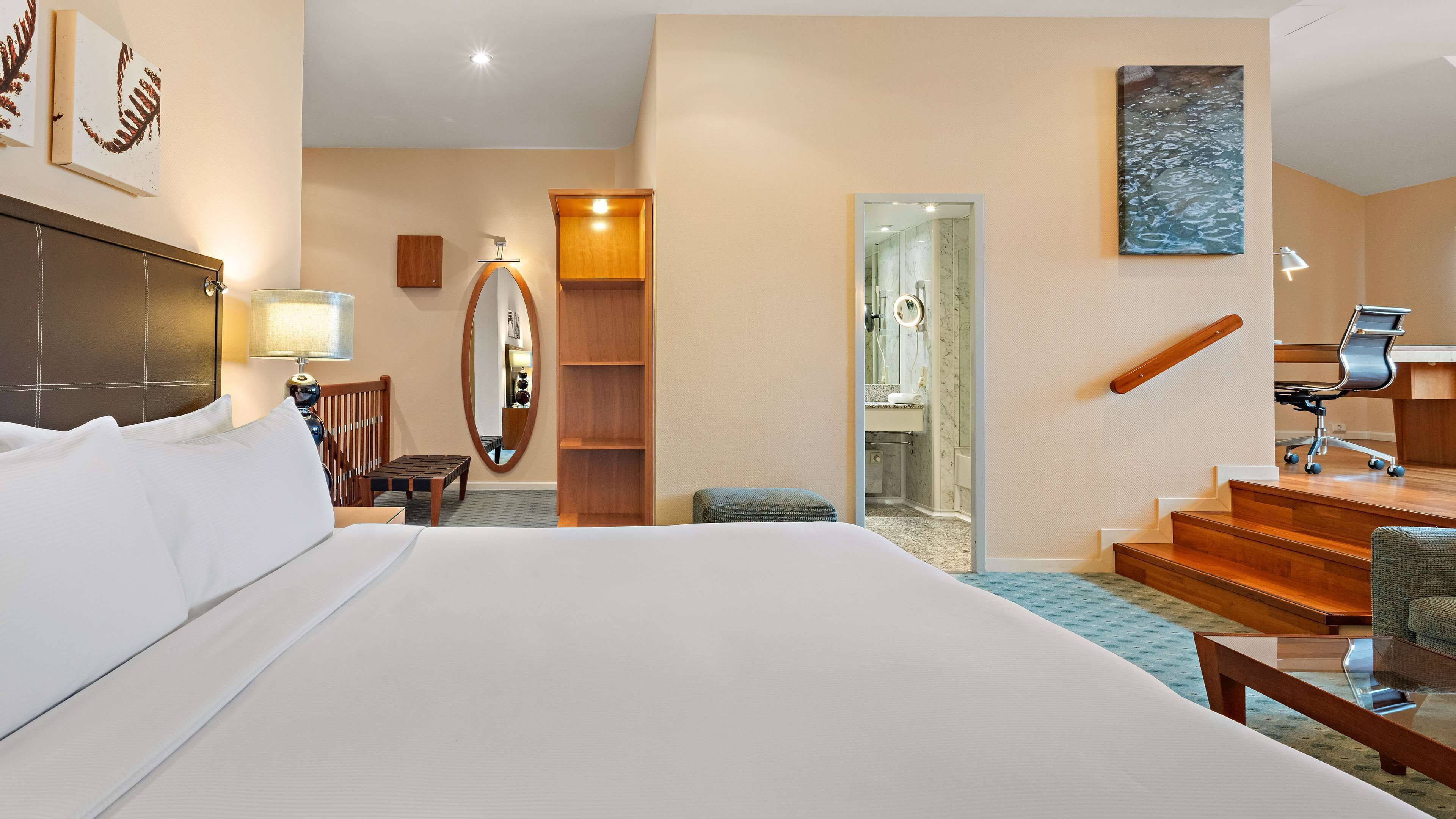 A hotel room with a large bed, a desk, a chair, a mirror, a wooden cabinet, and a staircase.