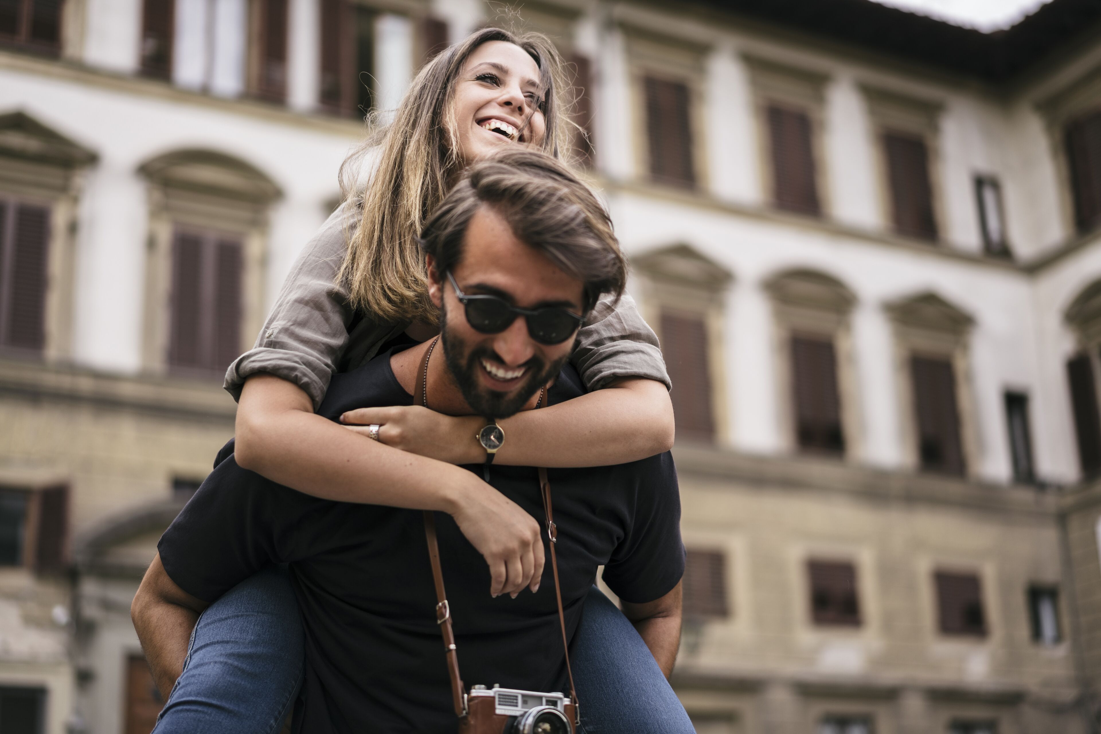 Un homme portant une femme sur son dos, avec un appareil photo suspendu à son cou, devant un bâtiment doté de fenêtres en arc de cercle.