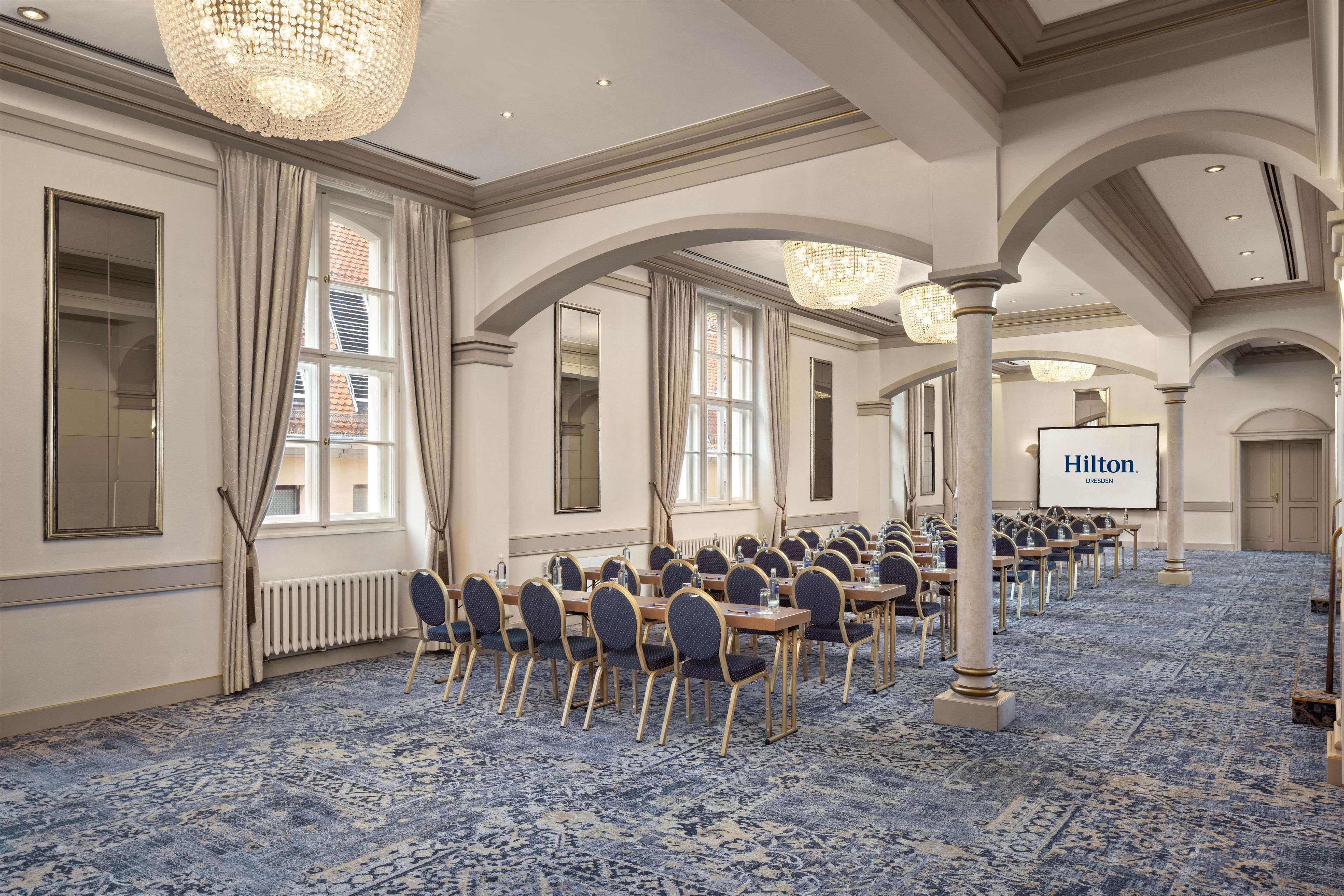 A hotel conference room with rows of tables and chairs, large windows, and chandeliers.