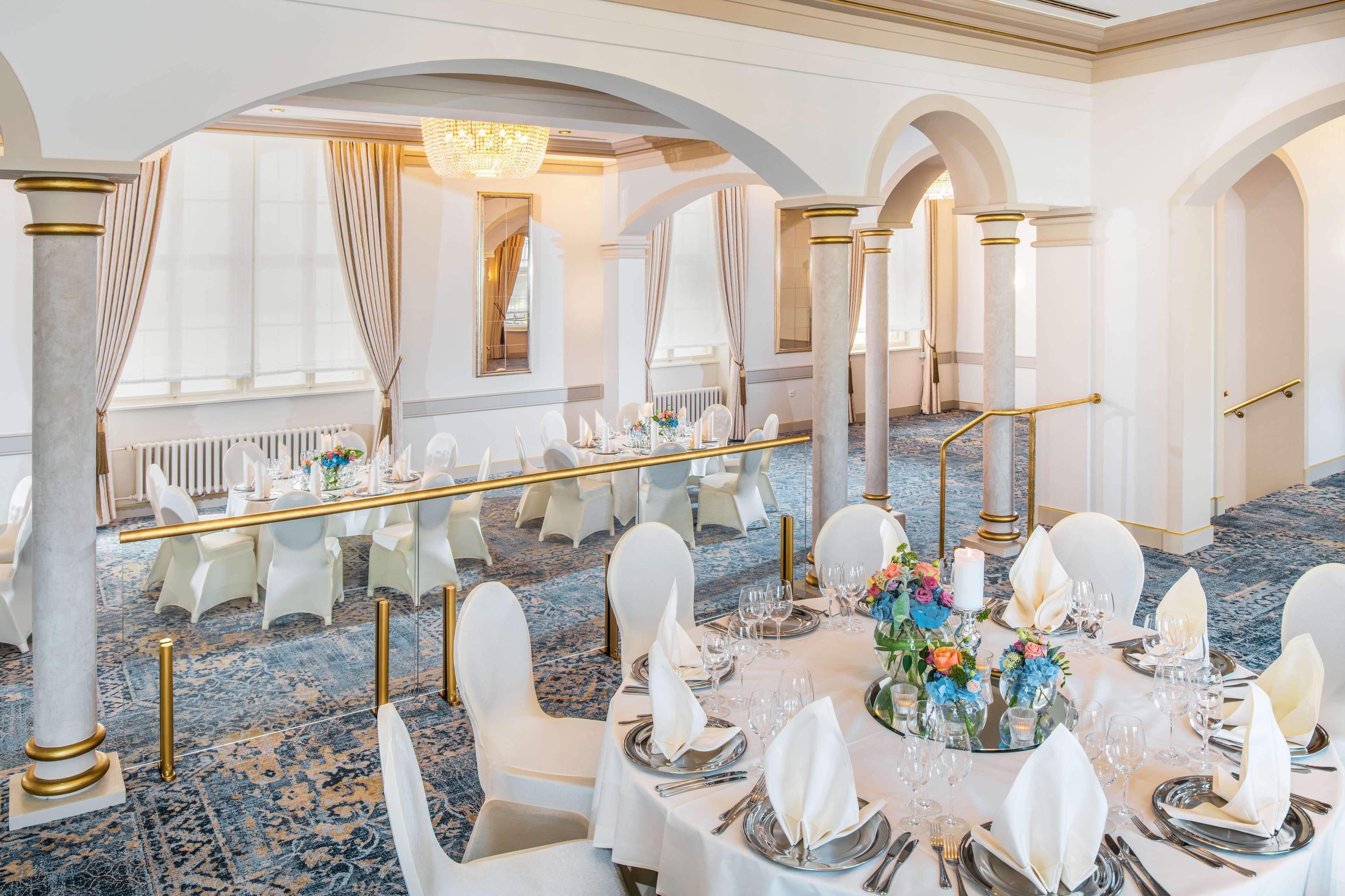 A banquet hall with round tables set for a formal event, featuring white chairs, gold railings, and a blue carpeted floor.