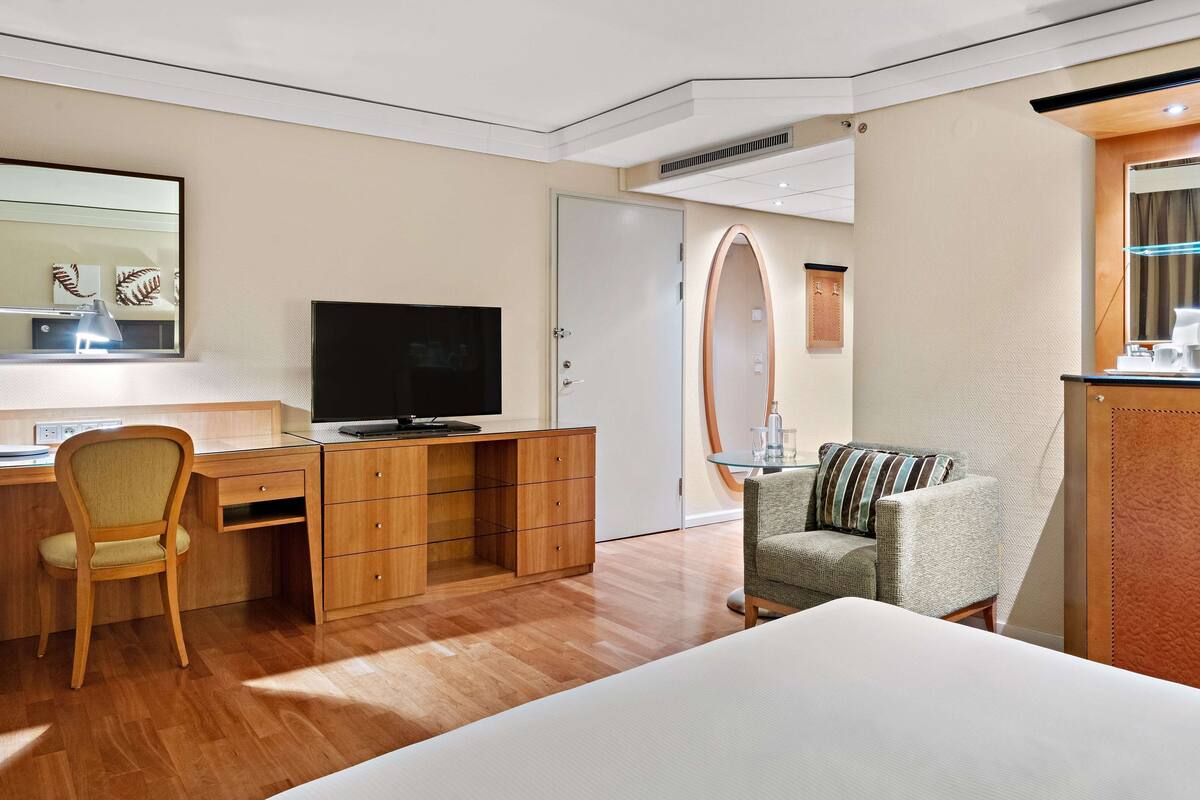 A hotel room with a wooden desk, a television, a bed, a chair, and a minibar.