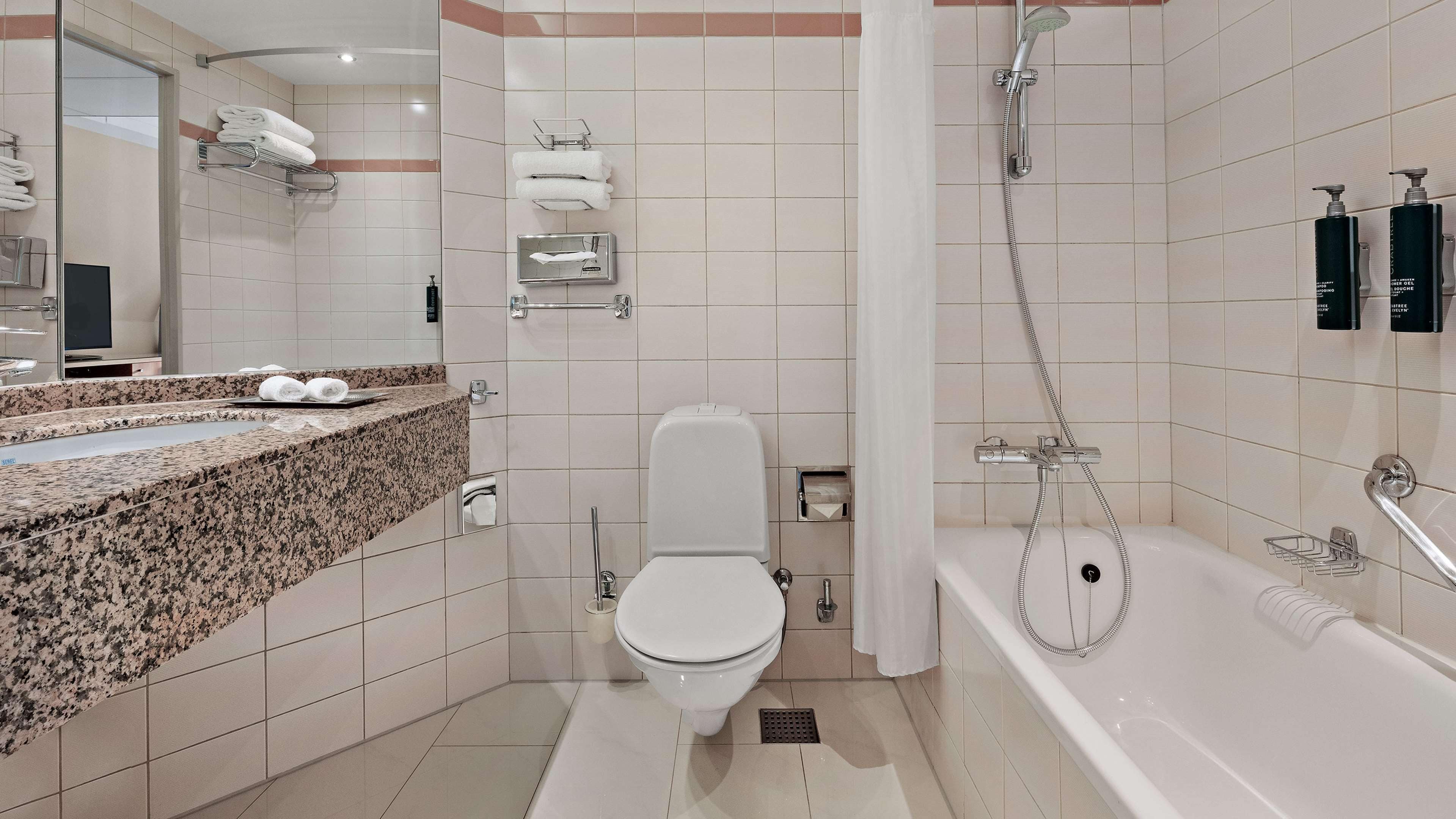 A modern bathroom with a granite countertop, a toilet, a shower with a handheld showerhead, and two bottles of shampoo on the wall.