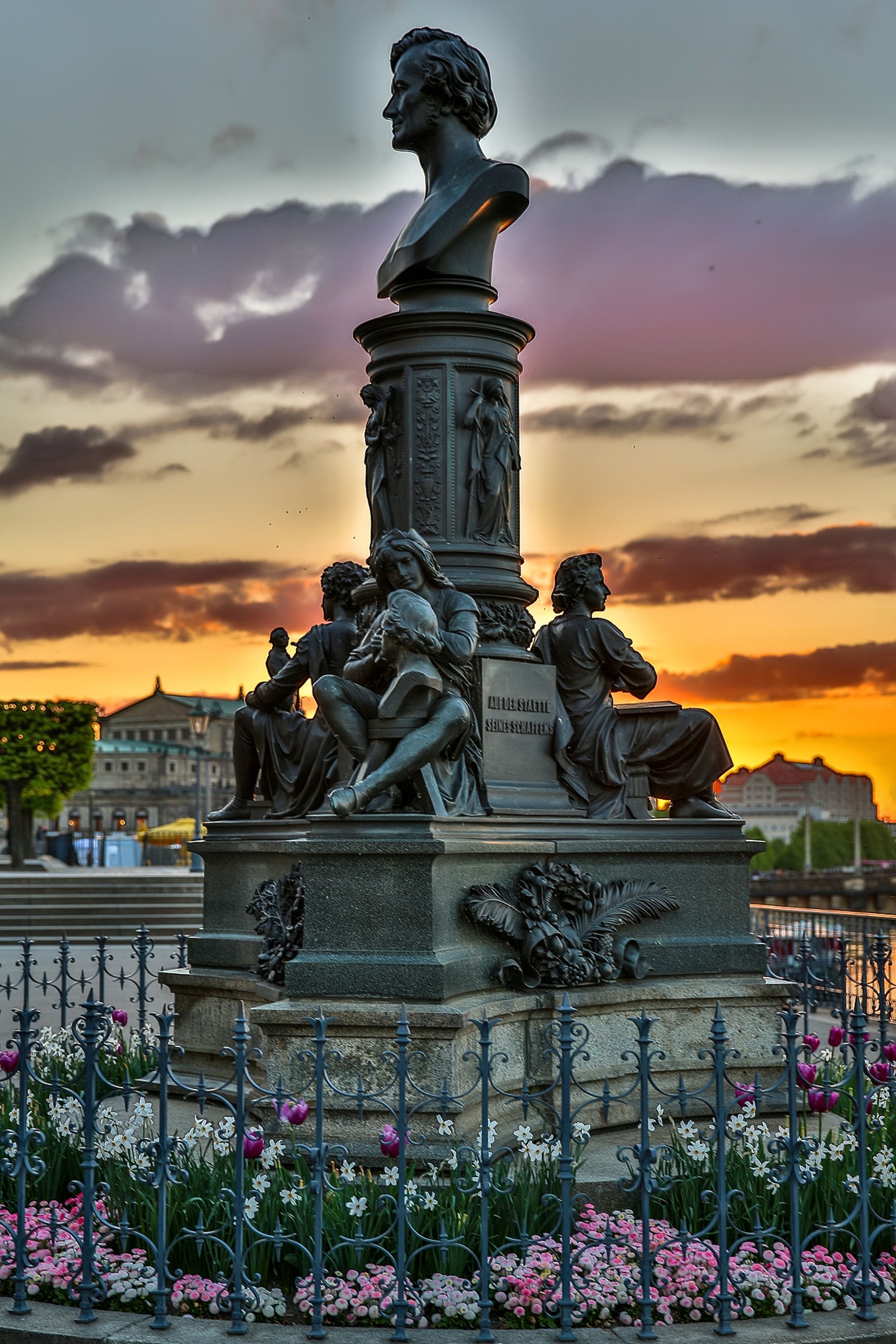Una escultura con figuras sentadas alrededor de un busto central, rodeada por una valla de metal y flores.