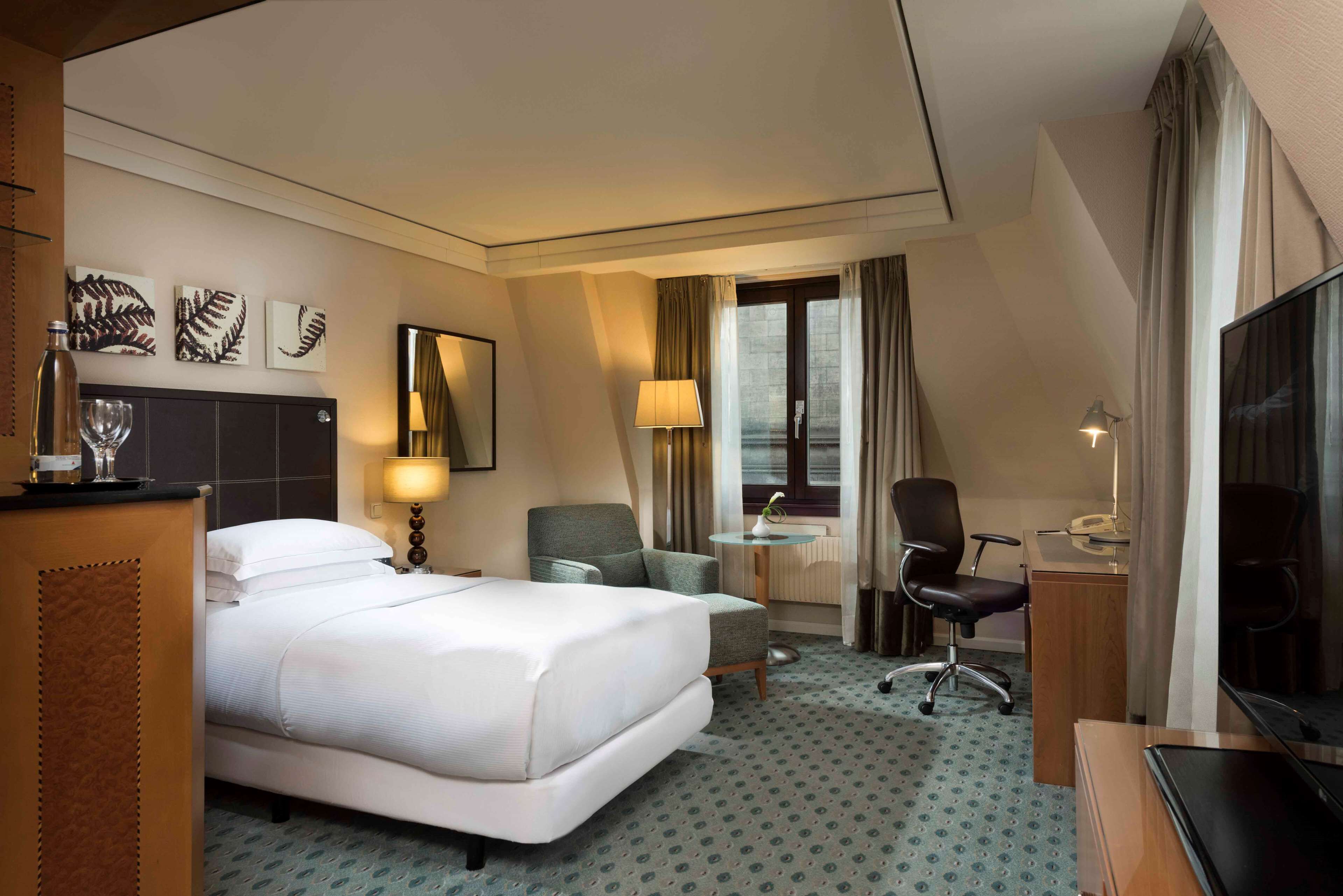 A hotel room with a bed, two chairs, a desk with a lamp, a television, and artwork on the wall.