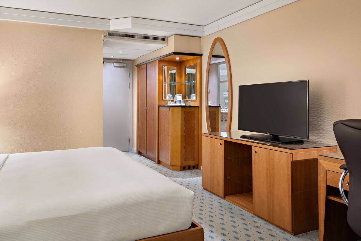 A hotel room with a bed, a desk with a television, a mirror, and a wardrobe.