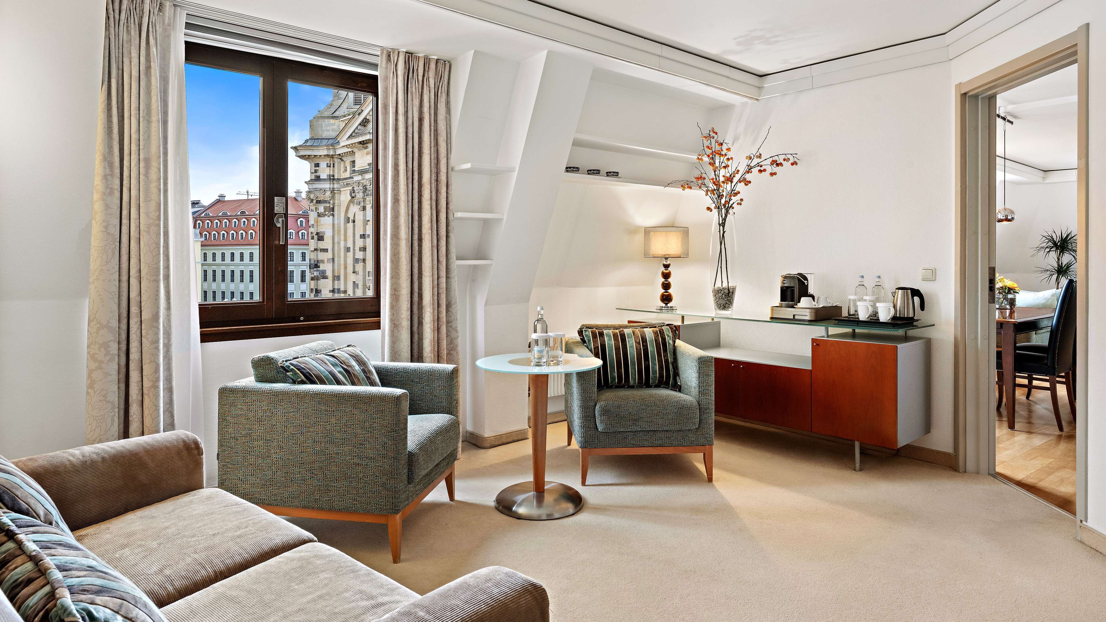 A modern hotel room with a sofa, armchairs, a small round table, a kitchen area, and a view of buildings through the window.