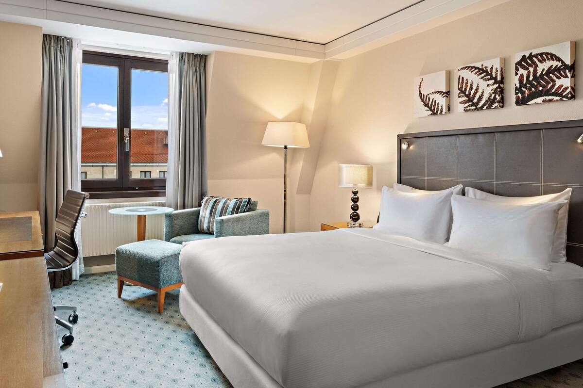 A hotel room with a large bed, a desk with a TV, a chair, a small table, and a window with curtains.