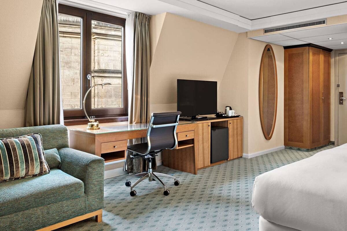 A hotel room with a bed, a desk with a computer, a chair, a sofa, a television, and a wardrobe.