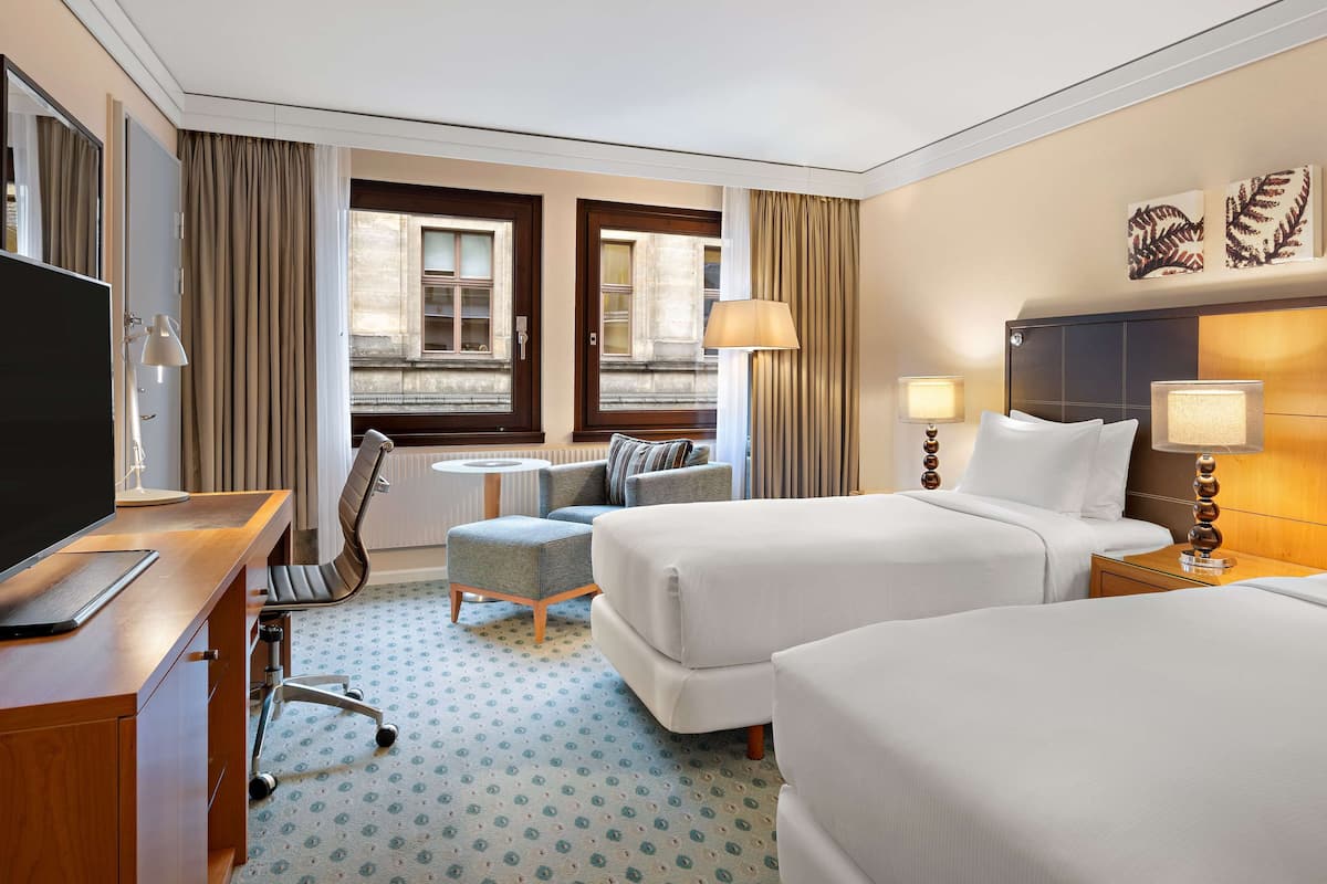 A hotel room with two beds, a desk with a TV, a small round table with a chair, and a window with curtains.