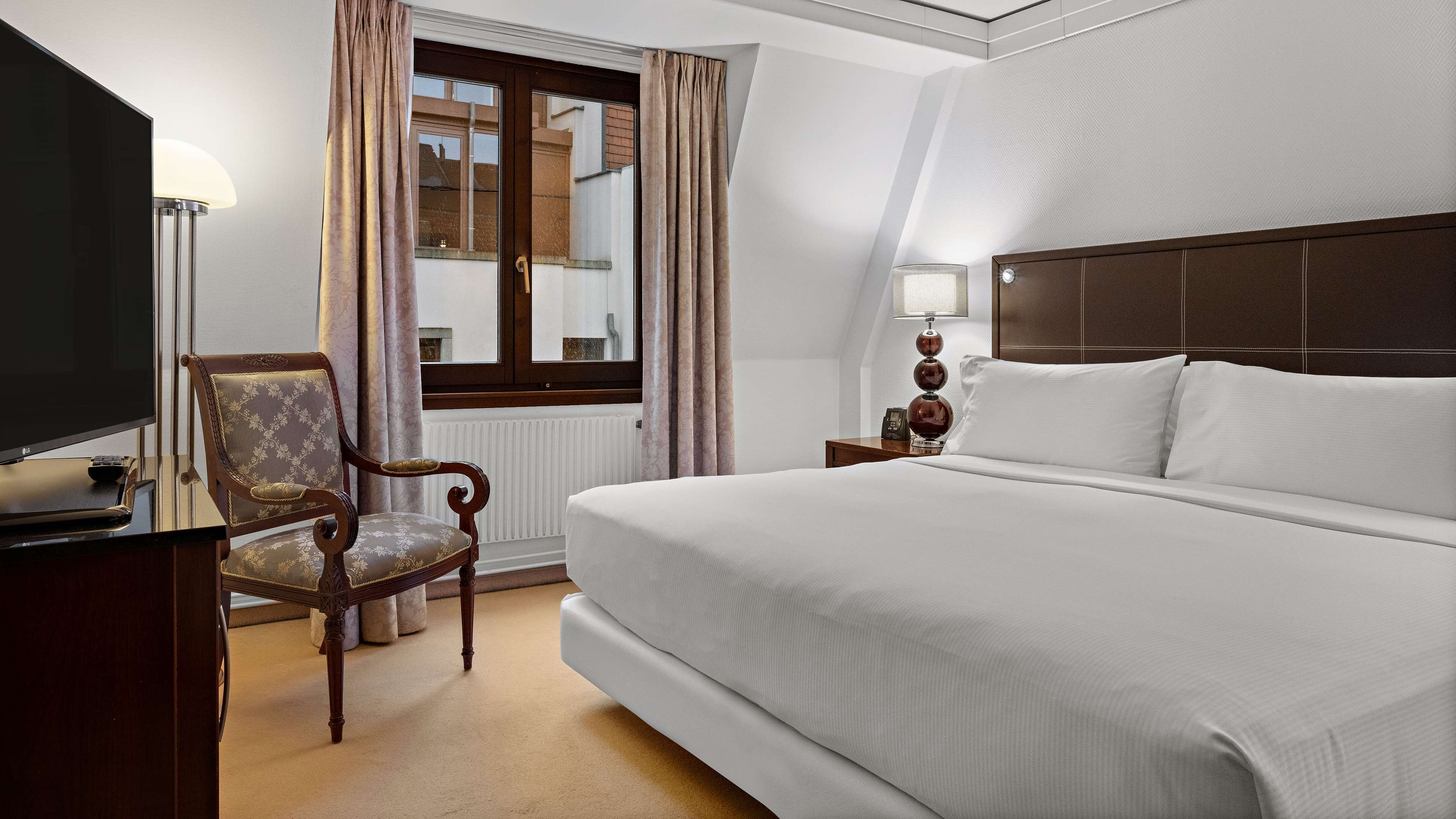 A hotel room with a large bed, a TV, a chair, a window with curtains, and a bedside table with a lamp.