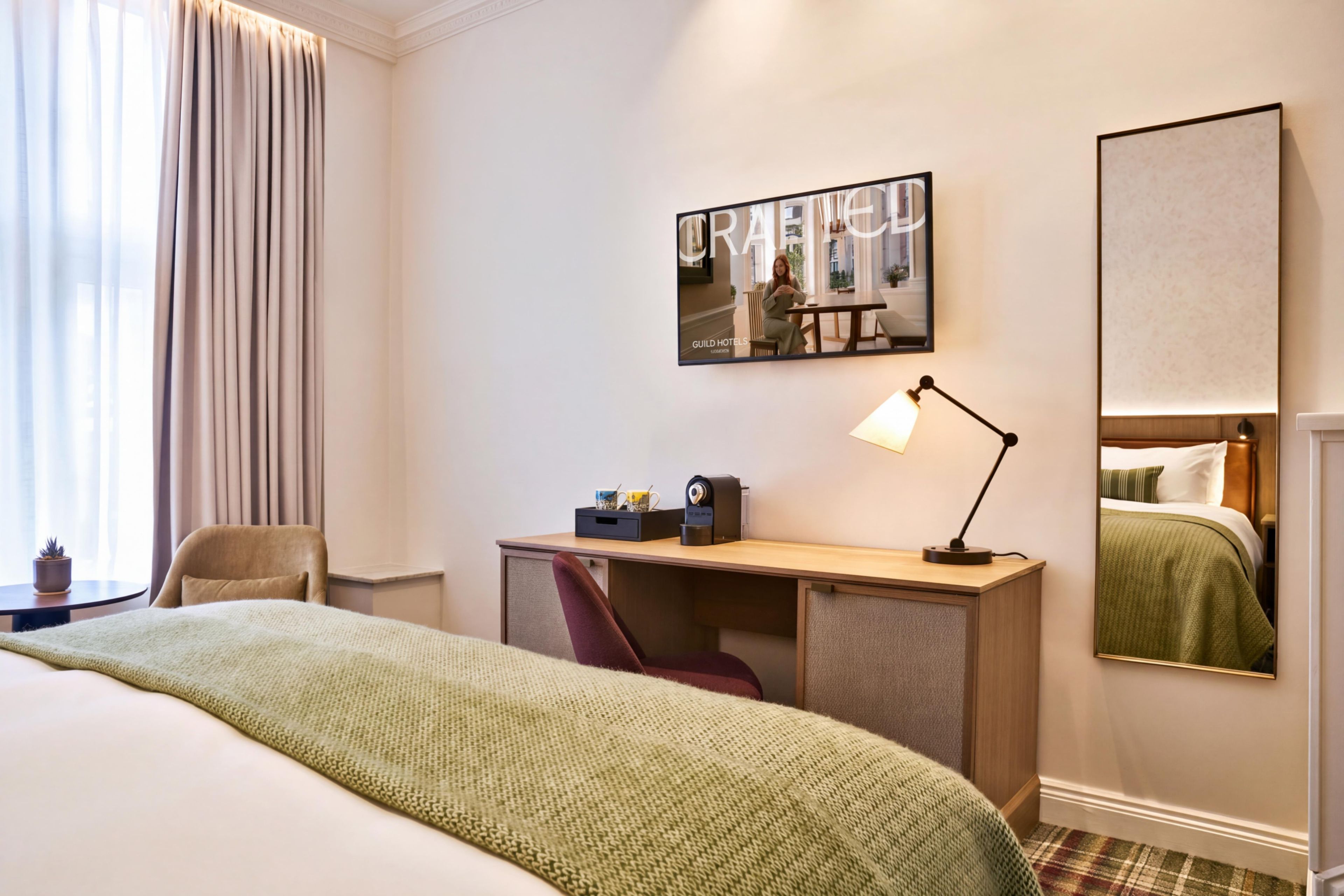 A hotel room with a bed, a desk, a chair, a lamp, and a large mirror.