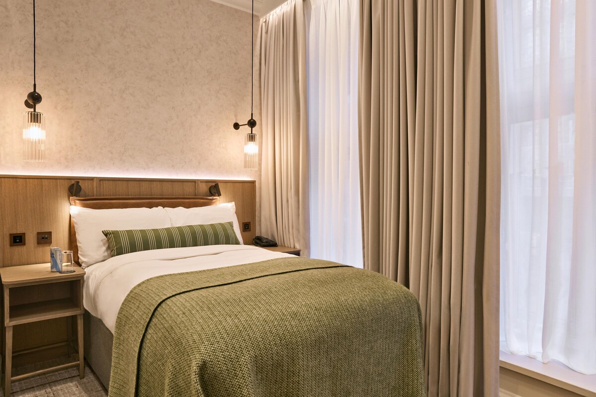 A hotel room with a bed, nightstands, a lamp, and a window with curtains.