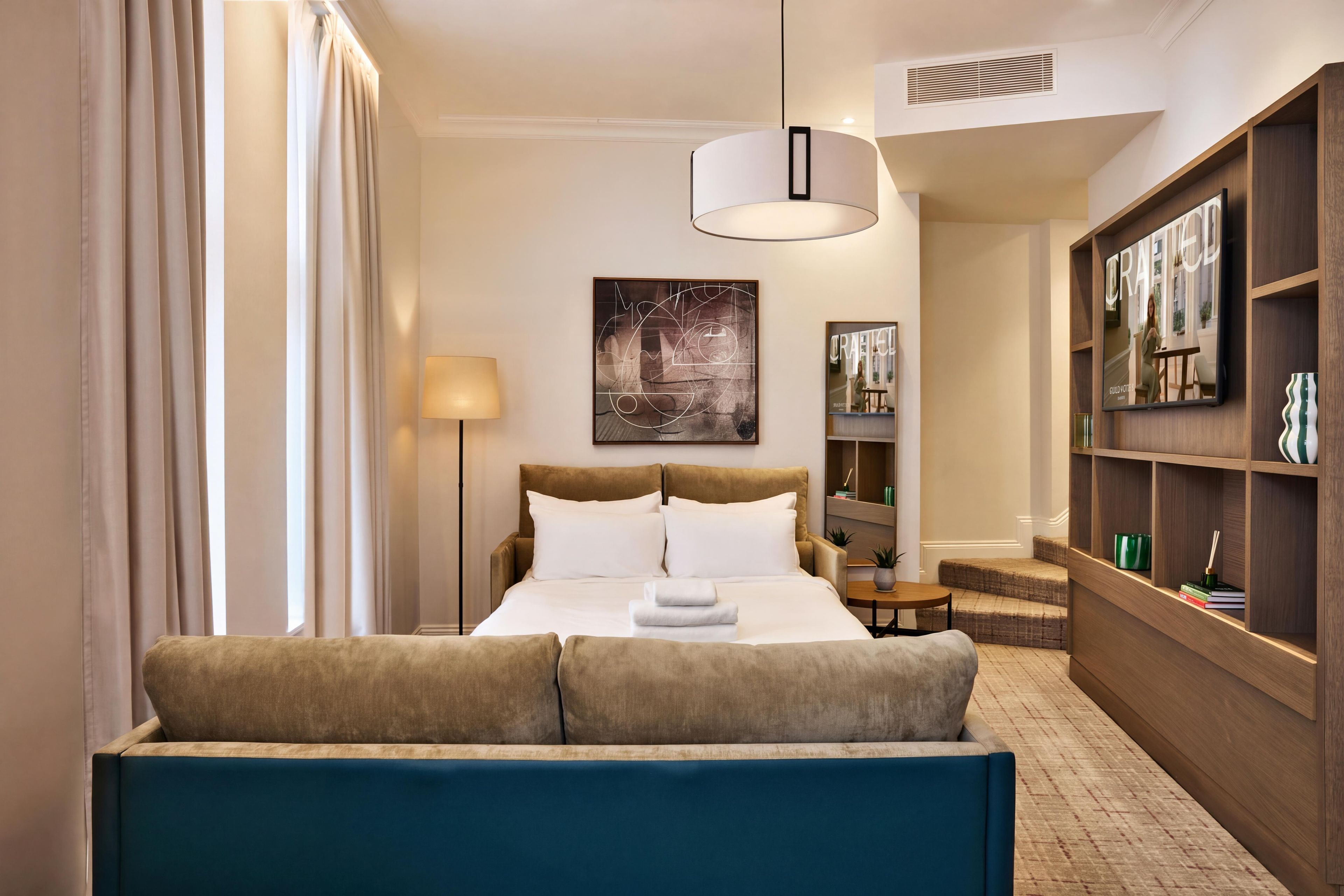 Una habitación de hotel con una cama, un sofá, una lámpara, un cuadro, una estantería y un televisor.
