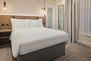 A neatly made bed with white linens, a wooden headboard, and a nightstand with a lamp.