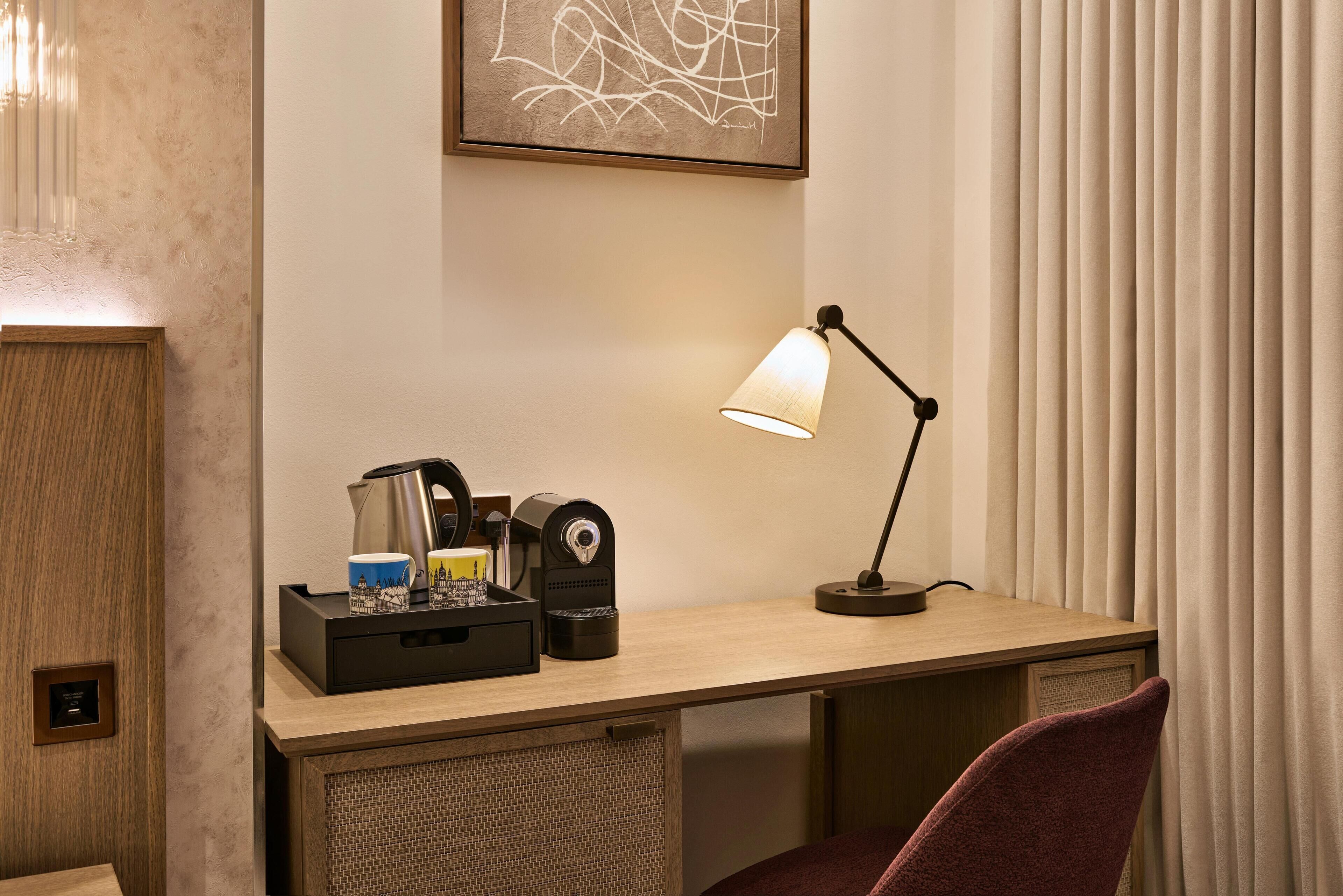 Habitación de hotel con un escritorio de madera, una cafetera, un hervidor, una taza y una lámpara.
