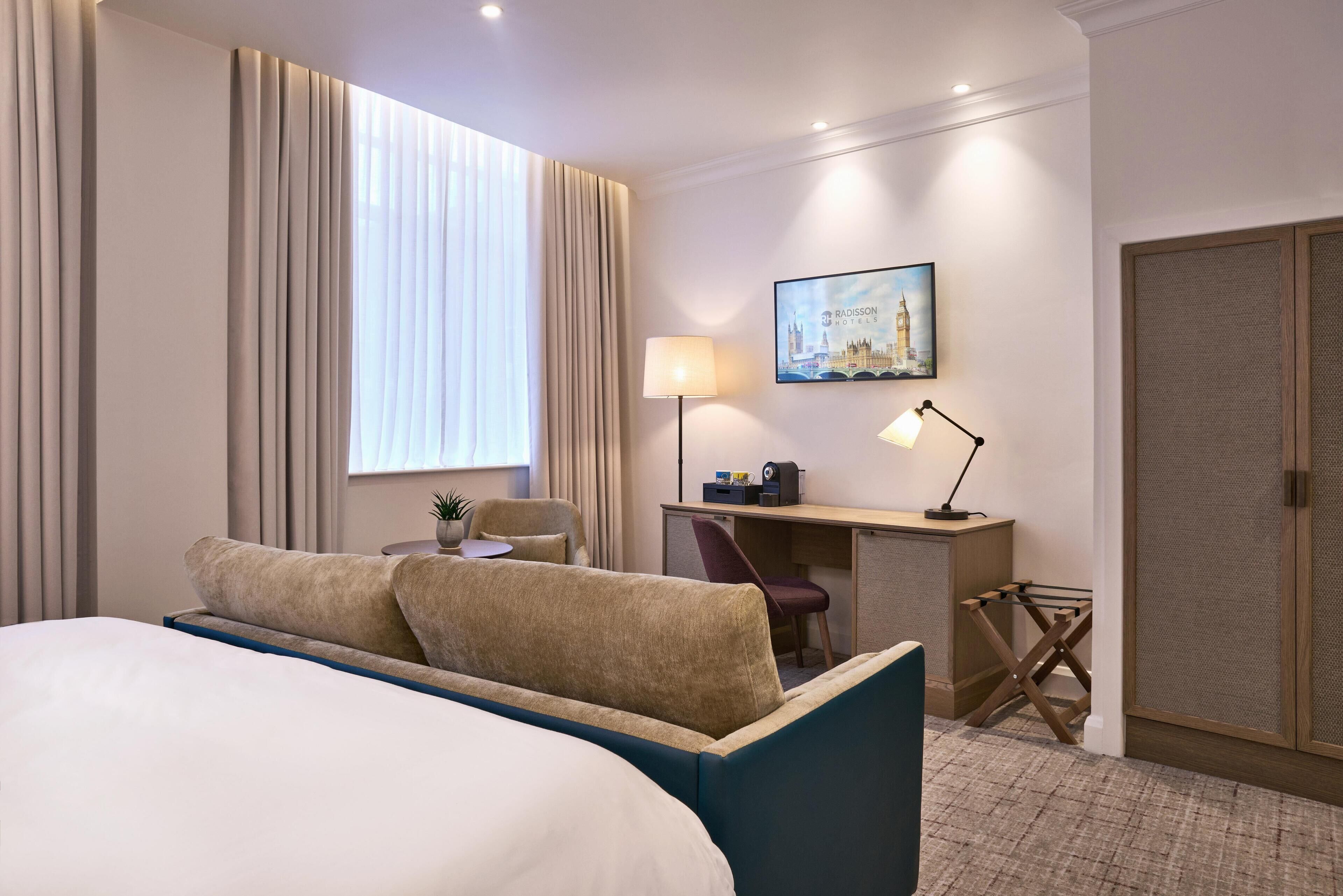 A hotel room with a bed, a desk, a chair, a lamp, and a painting of a cityscape.