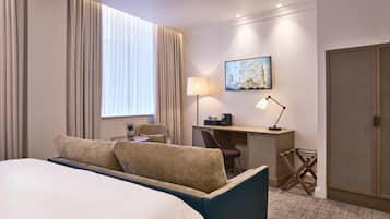 A hotel room with a bed, a desk, a chair, a lamp, and a painting of a cityscape.