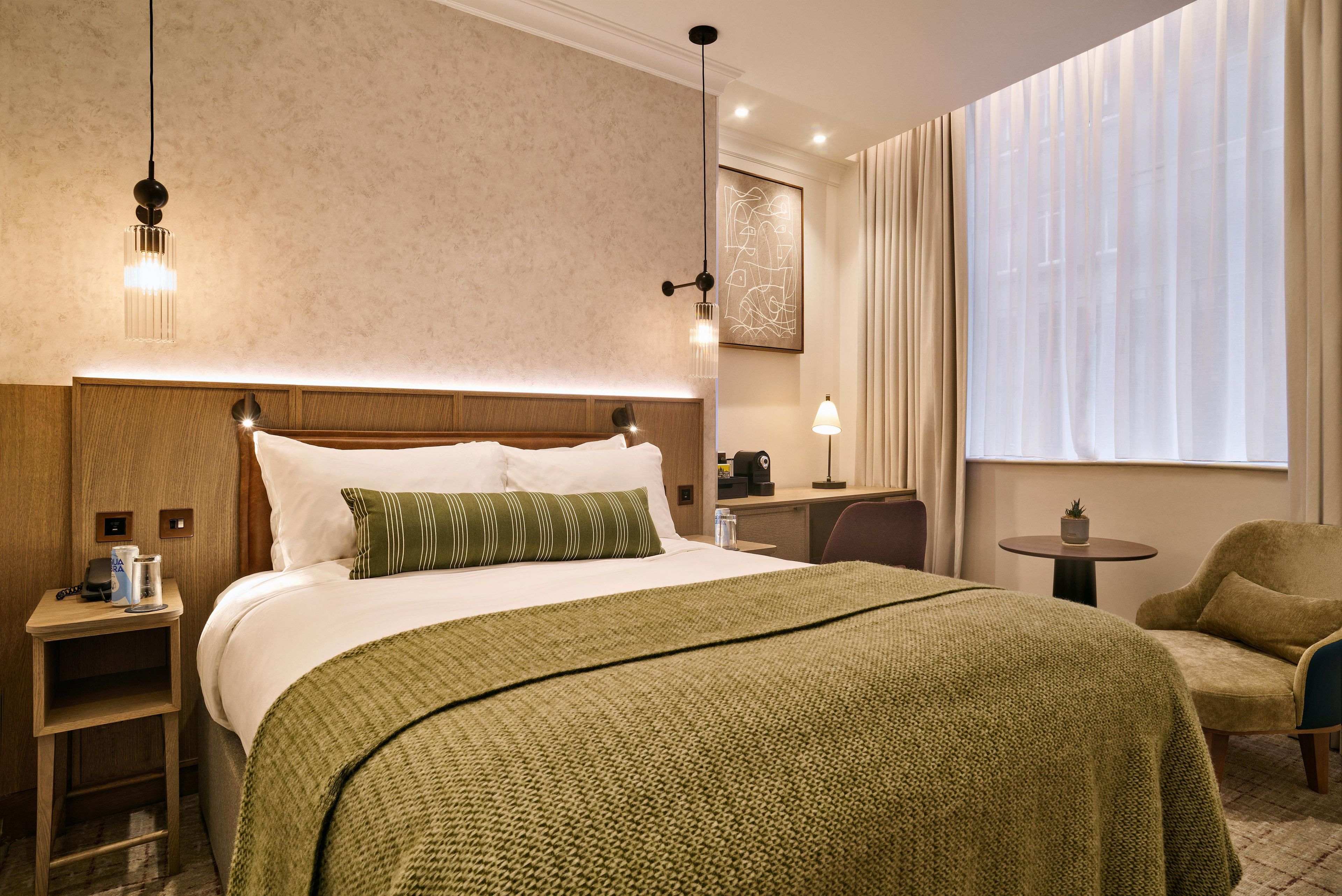 A hotel room with a large bed, bedside tables, a desk, a chair, a window with curtains, and a painting on the wall.