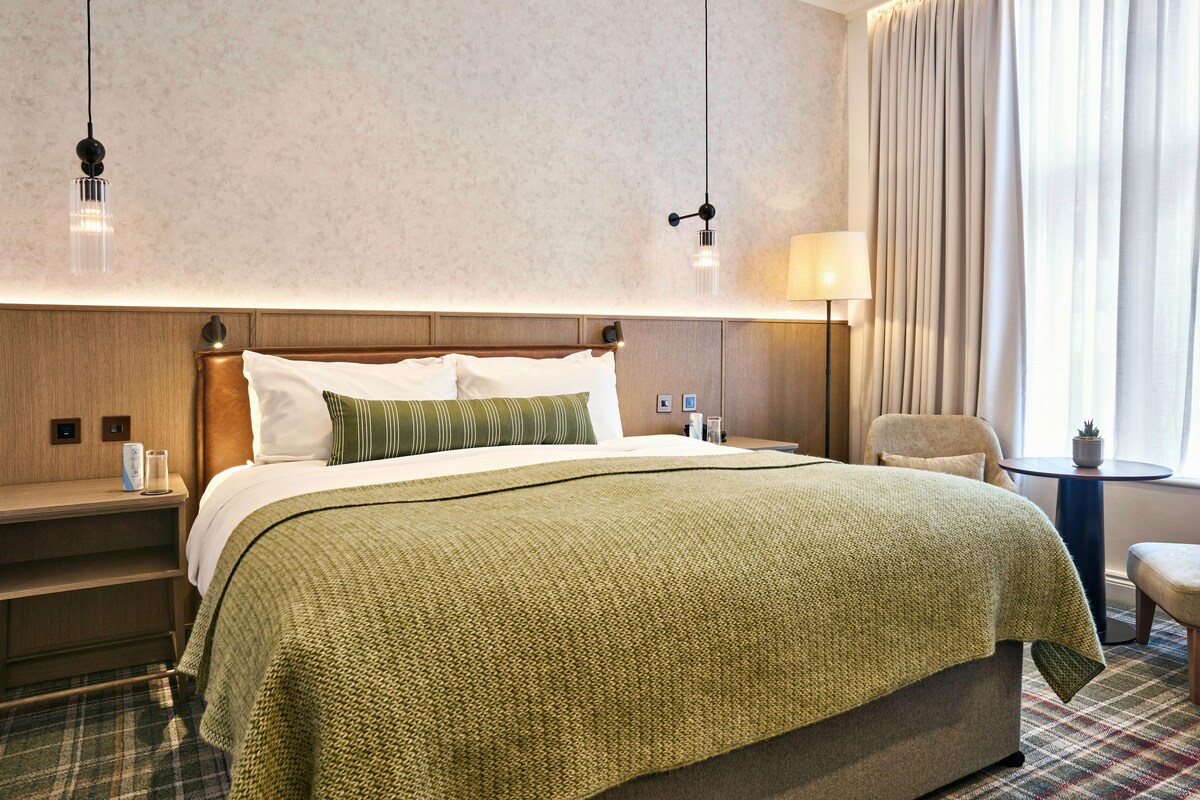A hotel room with a large bed, a bedside table, a lamp, a chair, and a window with curtains.