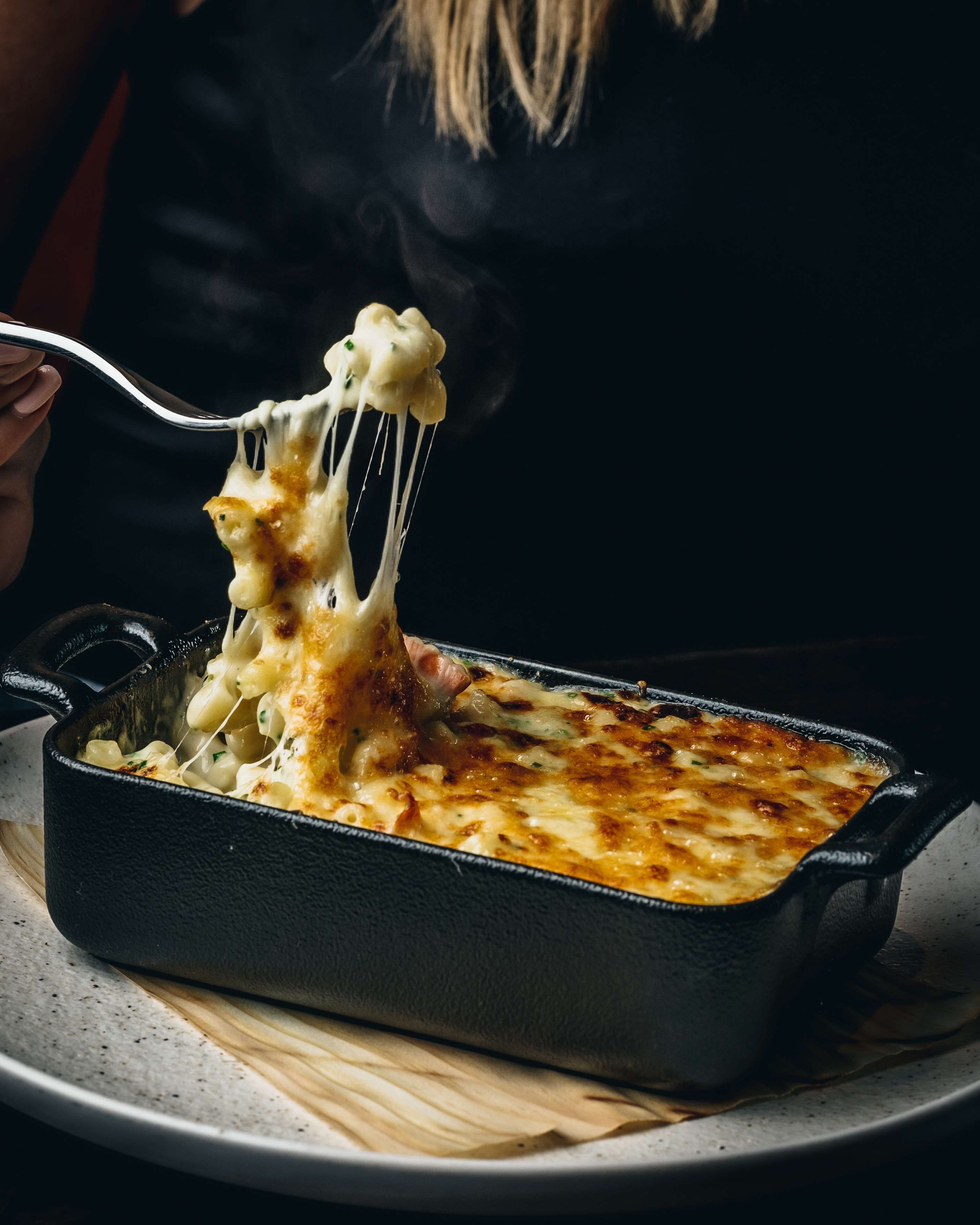 A person lifting a piece of macaroni and cheese from a black baking dish.