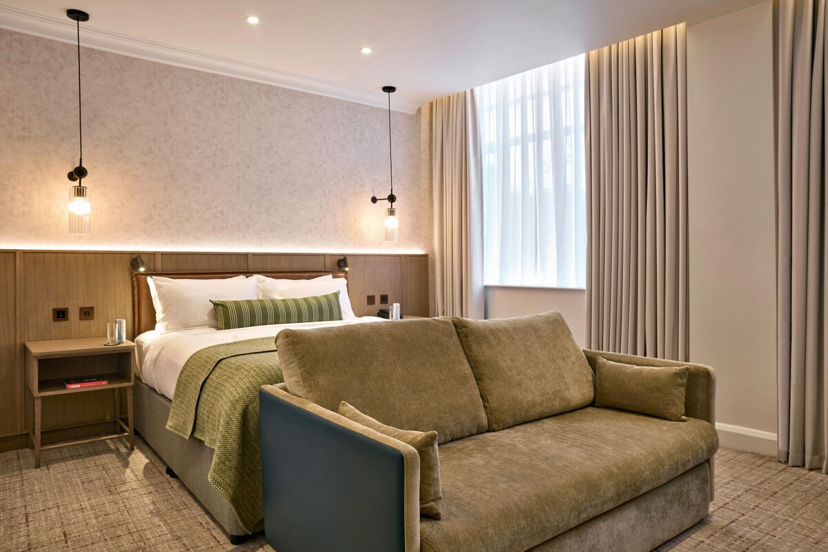 A hotel room with a bed, bedside tables, a sofa, a window with curtains, and pendant lighting.