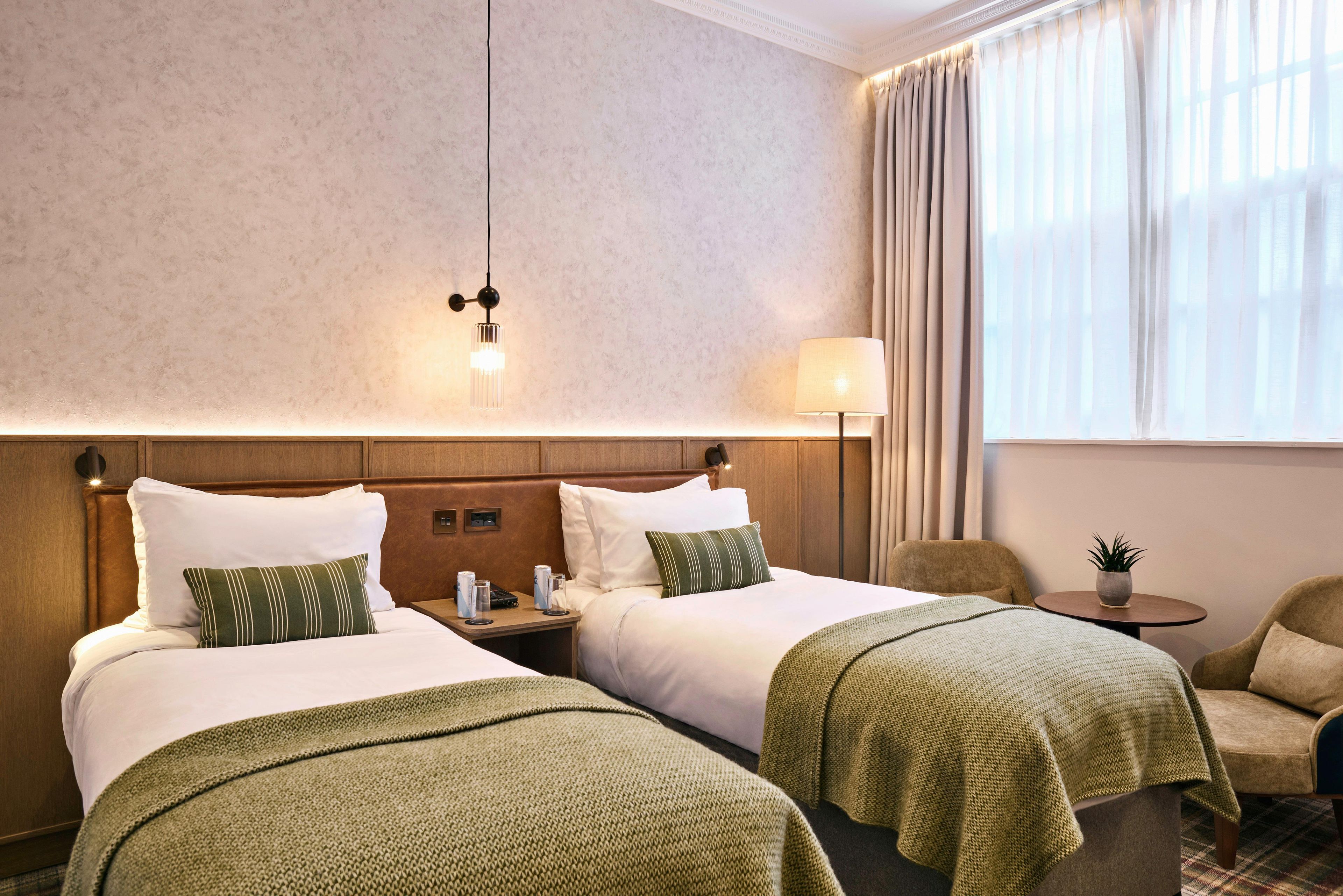 A hotel room with two beds, a nightstand, a lamp, a chair, and a small table with a plant.