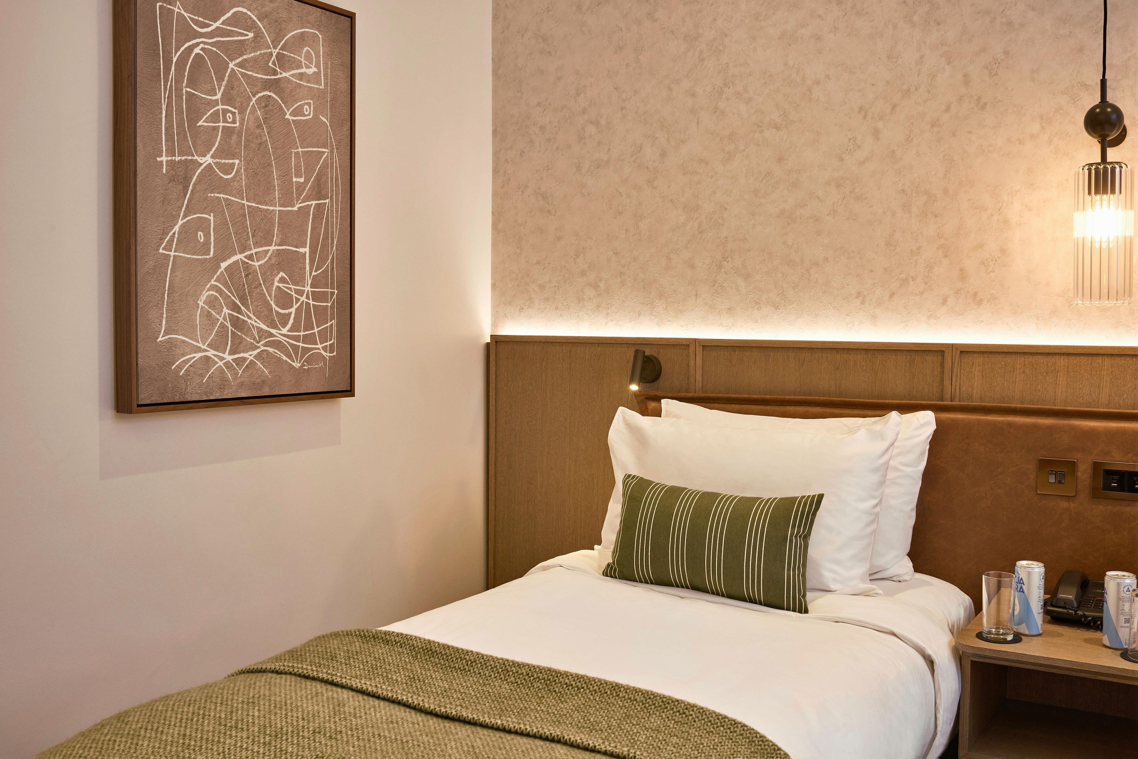 A hotel room with a bed, a nightstand, a lamp, a wall art piece, and a textured wall.