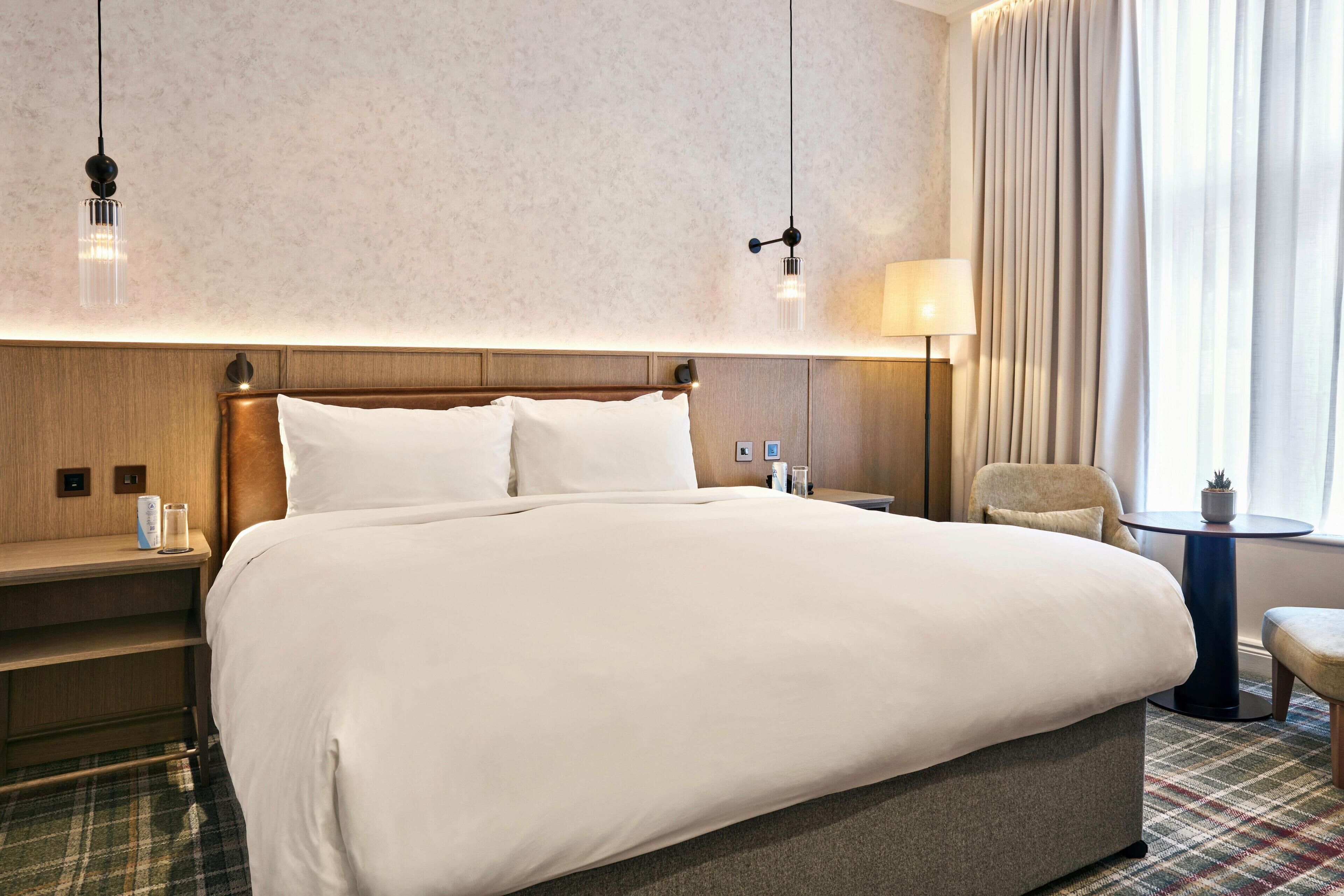 Une chambre d’hôtel avec un grand lit, une tête de lit en bois, une table de chevet avec une lampe, une chaise et une petite table avec une plante.