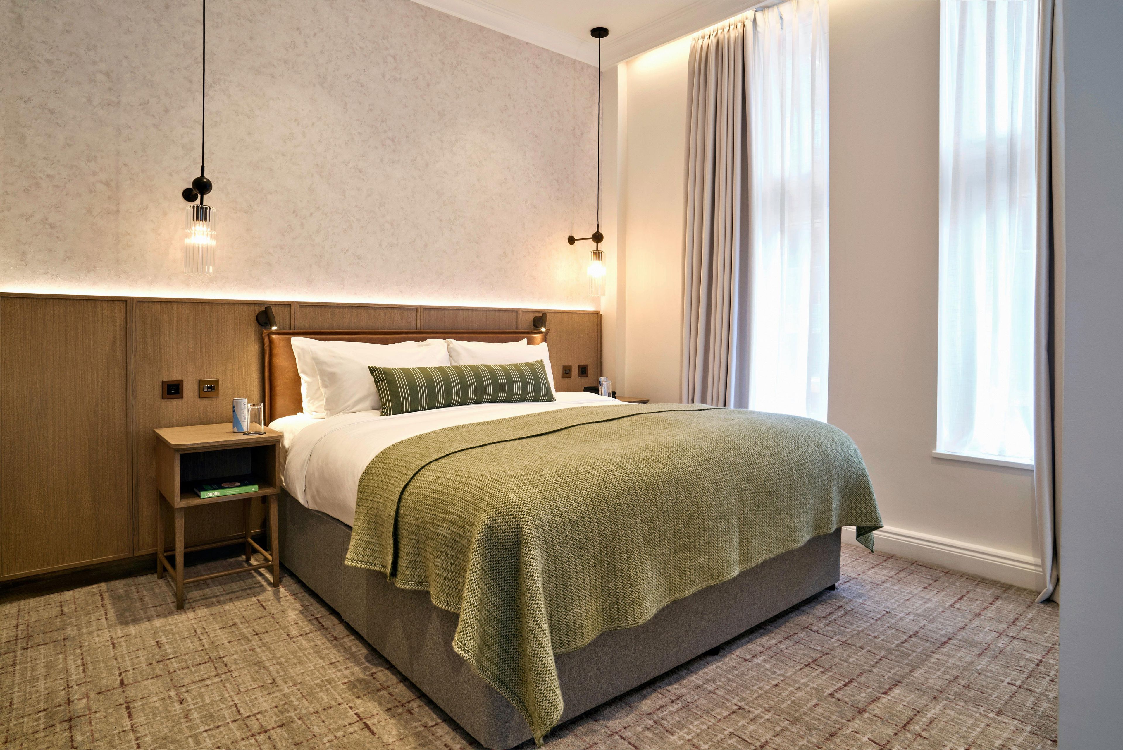 Una habitación de hotel moderna con una cama grande, una mesita de noche, una lámpara y un ventanal con cortinas.