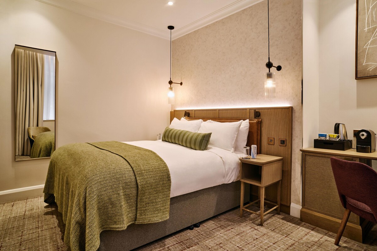 A hotel room with a bed, bedside table, lamp, chair, and a mirror.