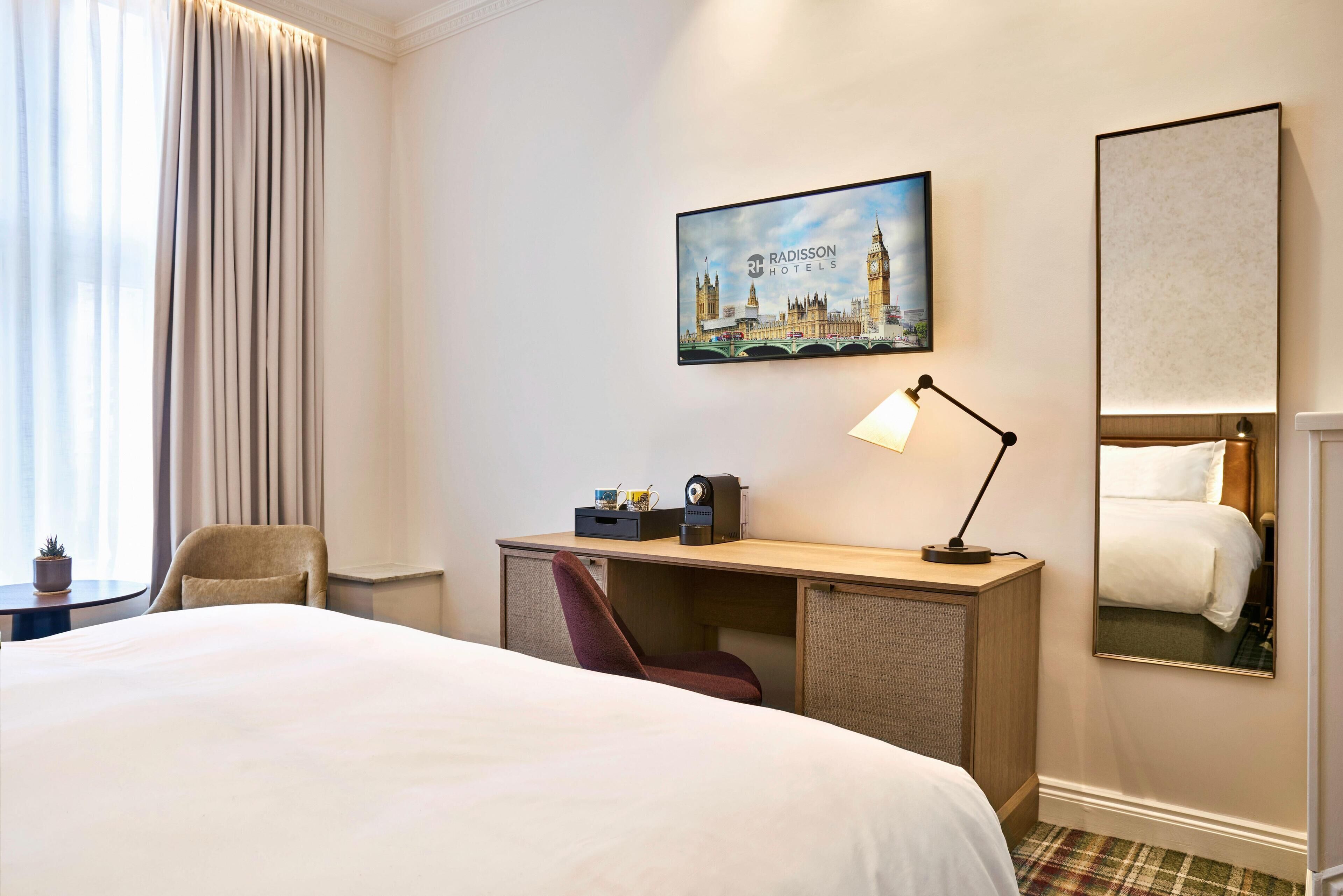 A hotel room with a bed, a desk with a lamp, a chair, a coffee maker, and a painting of a cityscape.