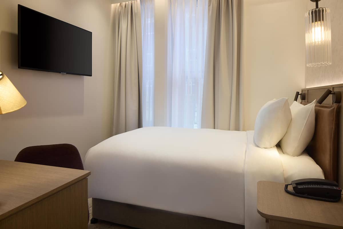 Habitación de hotel con televisor de pantalla plana en la pared, cama con ropa blanca, escritorio con lámpara y mesita de noche con teléfono.