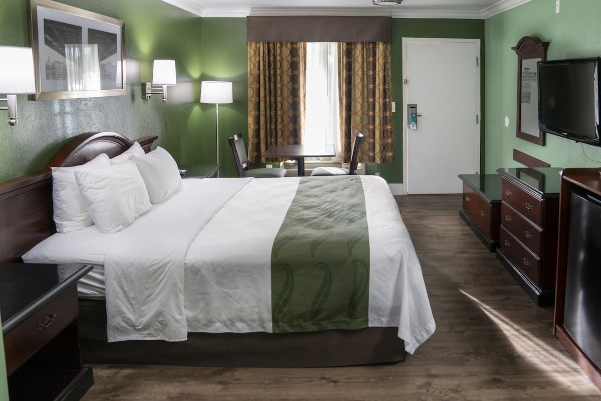 A hotel room with a large bed, a desk, a dresser, a television, and a window with curtains.